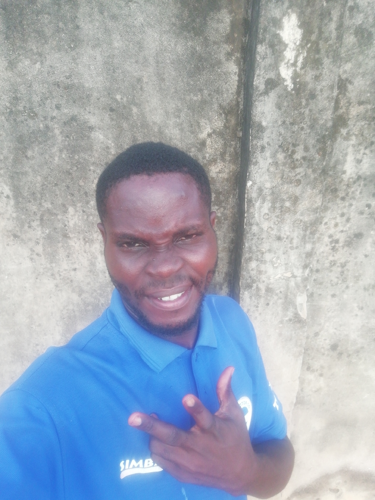 A dark-skinned man is taking a selfie outdoors, appearing close to a concrete wall with vertical and horizontal lines. He is wearing a blue polo shirt with "SIMB" printed on it. He is making a hand gesture with his left hand, showing his middle finger and index finger, while smiling. The location is the city of Unknown, Unknown.