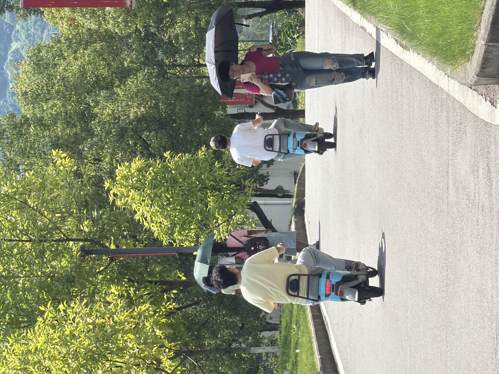 The image captures a bright, sunny day on a paved pathway, likely within a park or university campus in Jiangjin, China. Lush green trees line the path, casting dappled shadows. In the foreground, a person wearing a yellow t-shirt and blue jeans rides a light blue electric scooter. Further down the path, another person on a similar electric scooter, wearing a white t-shirt and a cap, is seen from behind. To the right, a person in a pink t-shirt and ripped blue jeans walks with a black umbrella open, holding a pink smartphone and a patterned bag. Another individual with a green umbrella and a backpack is visible behind the person on the first scooter. The ground is a smooth asphalt surface with a concrete curb and grassy areas on either side. The overall atmosphere suggests a leisurely outdoor activity.