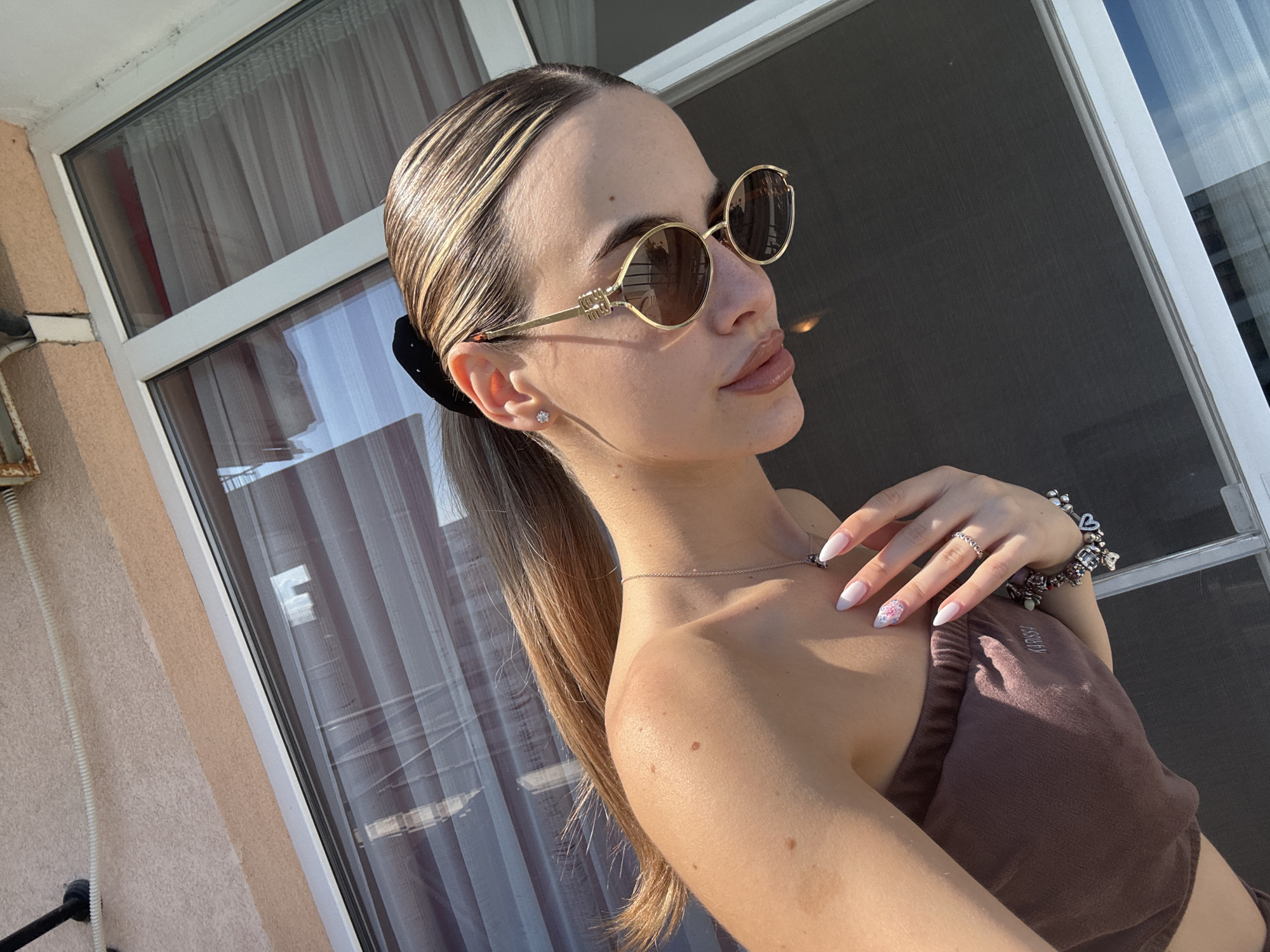In an outdoor setting, a young woman with blonde hair pulled back in a ponytail poses for a selfie. She is wearing gold-rimmed oval sunglasses, a simple silver necklace, and a brown top. Her hand, adorned with a silver ring and a bracelet with multiple charms, is resting on her shoulder. Her nails are painted white with a floral design on one finger. The background consists of a glass door with sheer curtains and a textured wall. The lighting suggests it is daytime, with sunlight casting shadows. This scene is likely located in Nesebar, Bulgaria, given the architectural style of the building visible in the background.