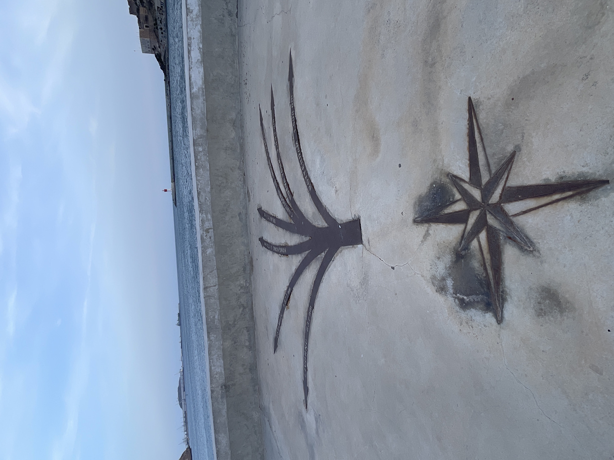 The image shows a concrete ground with two metallic engravings. In the foreground, a compass rose design is etched into the concrete. Behind it, a stylized tree-like shape branches out with each branch pointing towards a different city, with the city name and distance inscribed on it. The names visible include "ISTANBUL 2.537 Nm", "HONG KONG 9.171 Nm", "LIVORNO 937 Nm", "MADRID 322 Nm", "CARTAGENA DE INDIAS 7.330 Km", "NUEVA YORK 6.443 Km", and "ROTTERDAM 1.509 Nm". In the background, the sea is visible beyond a concrete barrier, with a lighthouse and a fortified building on a hill to the right, indicative of the city of Cartagena, Spain. The sky is cloudy with a light blue hue. There are no people visible in the image.