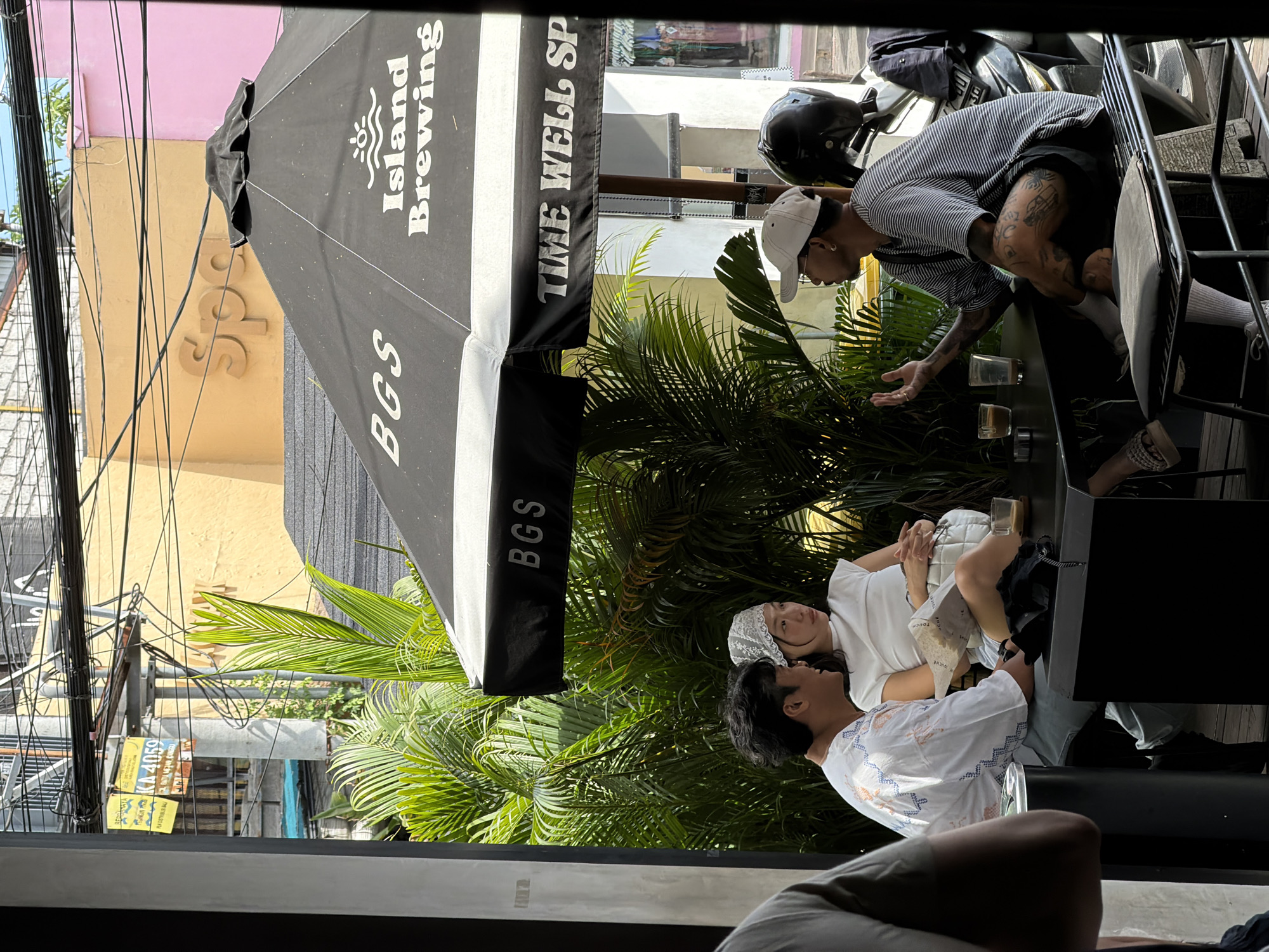 The image captures an outdoor scene, likely in North Kuta, Indonesia, as indicated by a local vehicle registration plate visible in the lower left. The foreground shows a partially visible person seated in what appears to be a lounge area, possibly inside a building or under a sheltered space.
The midground features an outdoor table surrounded by lush tropical foliage, likely palm trees. Four individuals are visible at the table. Two people sit opposite each other, one wearing a white patterned shirt, and the other wearing a white shirt and a lace head covering. Across from them, another person wearing a striped shirt with dark shorts stands and gestures towards the seated people, while a fourth person's legs are visible by the side of the table.
An umbrella with the branding "Island Brewing" and "TIME WELL SPENT" is positioned above the table, partially blocking the view. Electrical wires and utility poles are visible in the background, along with building facades painted in various colors.