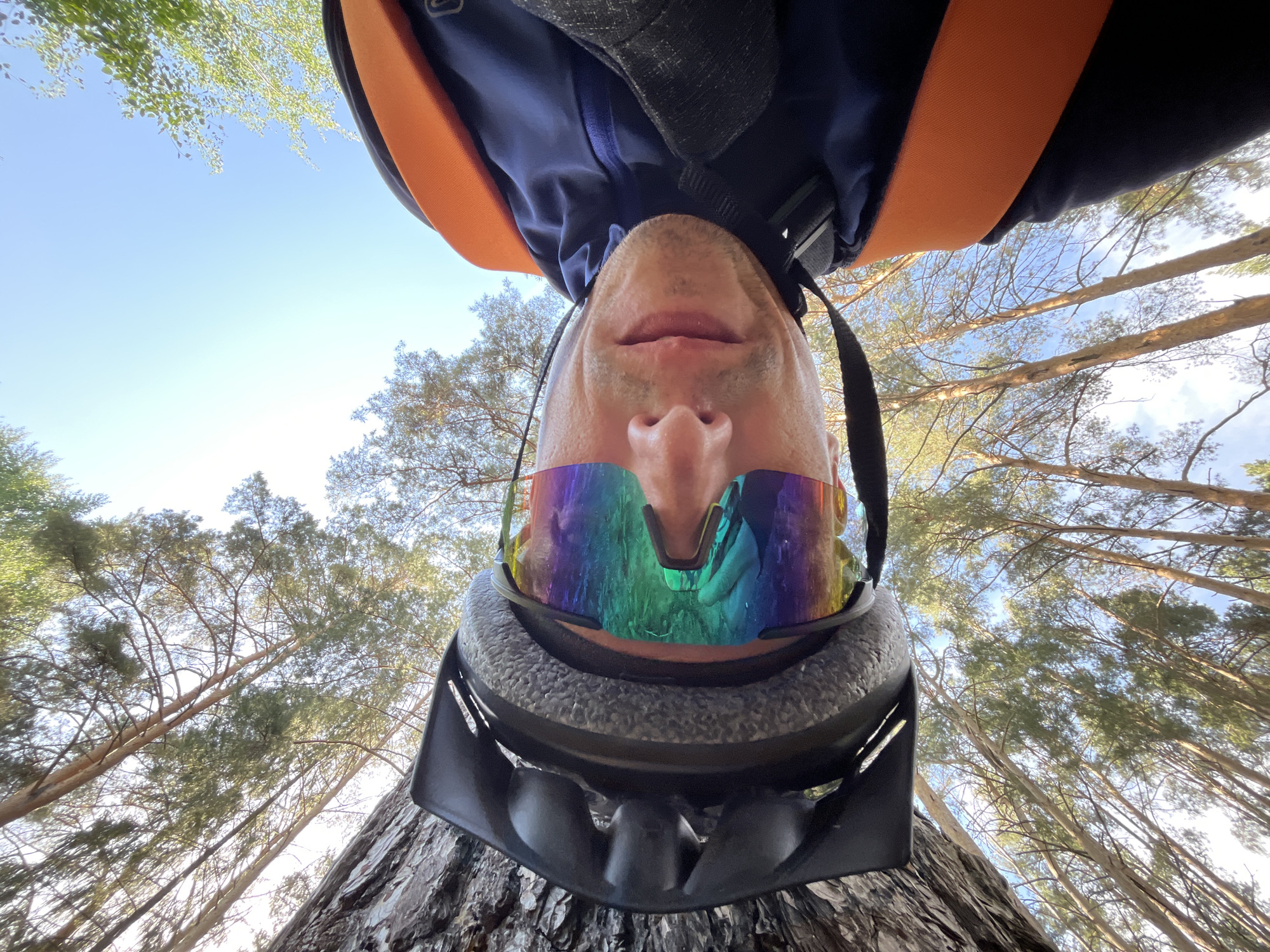A low-angle selfie photograph shows a person wearing a bicycle helmet and rainbow-tinted sports glasses looking up into a forest. The person is also wearing a dark blue jacket and a backpack with orange straps. The backdrop is a forest of tall trees with green foliage. The sky is visible in the upper portion of the frame. The photo was taken upside down. This photo may have been taken near Chaikovsky, Russia, due to being tagged with this location.