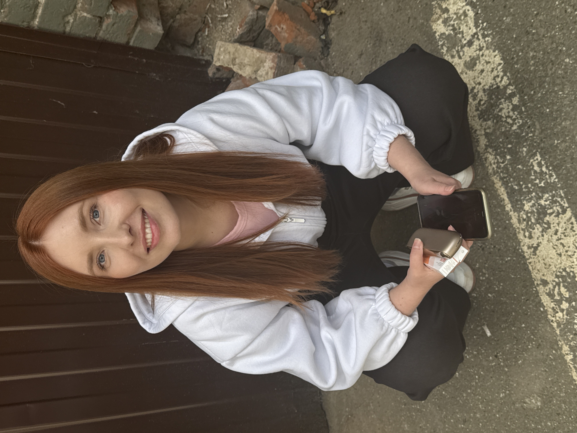 A young woman with long, reddish-brown hair and blue eyes is crouching outdoors. She is wearing a white zip-up hoodie over a pink t-shirt and black pants. She is holding a phone in her left hand and a small, rectangular object, possibly a vaping device, in her right hand. The background consists of a dark brown corrugated metal door on the left and a brick wall with scattered bricks on the right. The ground is asphalt with white markings. The scene appears to be in an urban environment, possibly an alleyway or a service area. There are no visible indications that the location is specifically in the city of Perm, Russia.