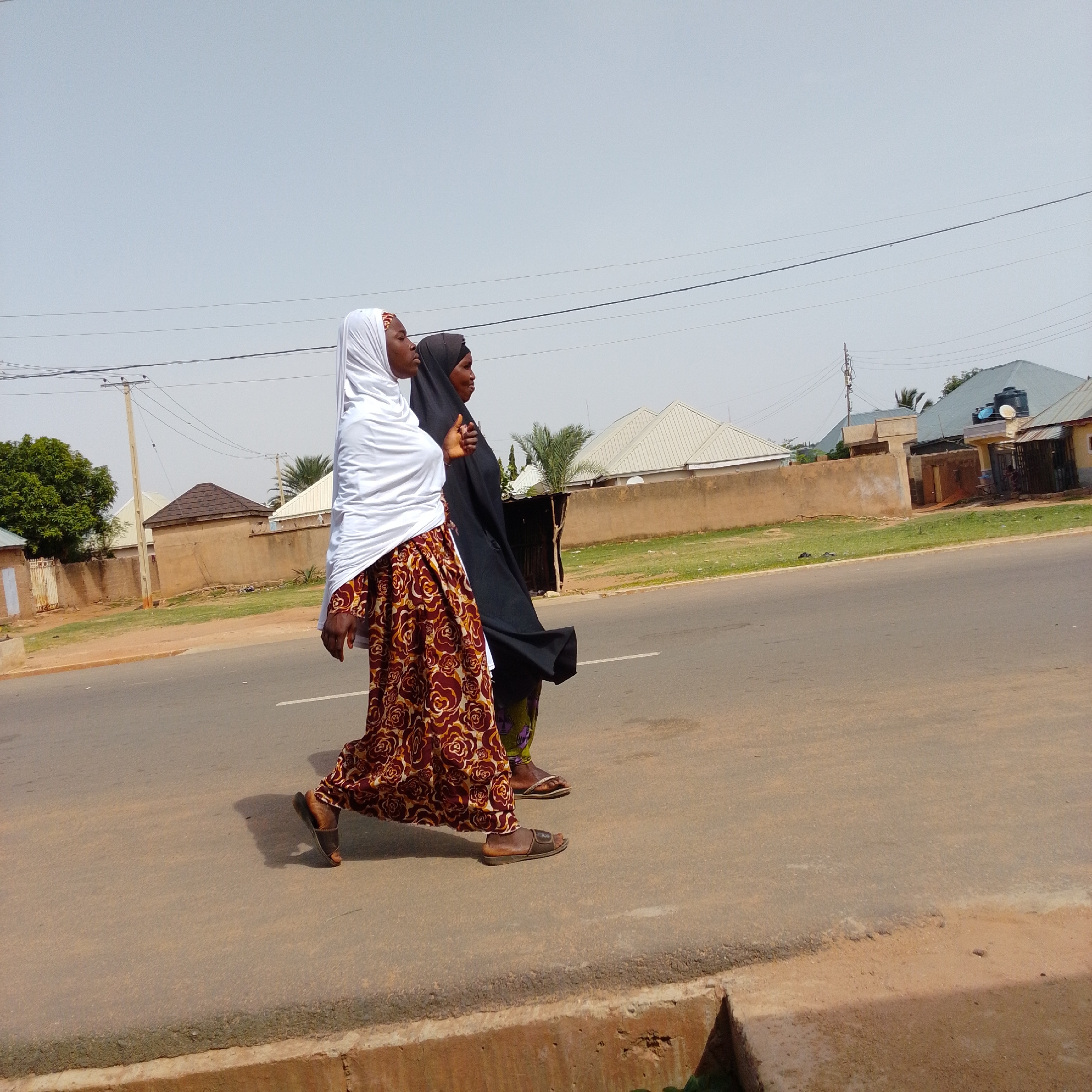 In Gombe, Nigeria, two individuals are depicted from the waist down, walking side-by-side on an asphalt road. The person on the left is wearing a white hijab and a long, patterned skirt with a maroon base and a floral design in shades of brown, yellow, and white. They are wearing brown sandals. The person on the right is wearing a black hijab and a patterned skirt with a green and purple design. They are also wearing sandals. The road is bordered by a concrete drain on the right. In the background, there is a beige wall, some green grass, and several buildings with light-colored roofs and some visible architectural details. Utility poles with wires are also present in the background. The sky is clear and light blue, suggesting daytime.