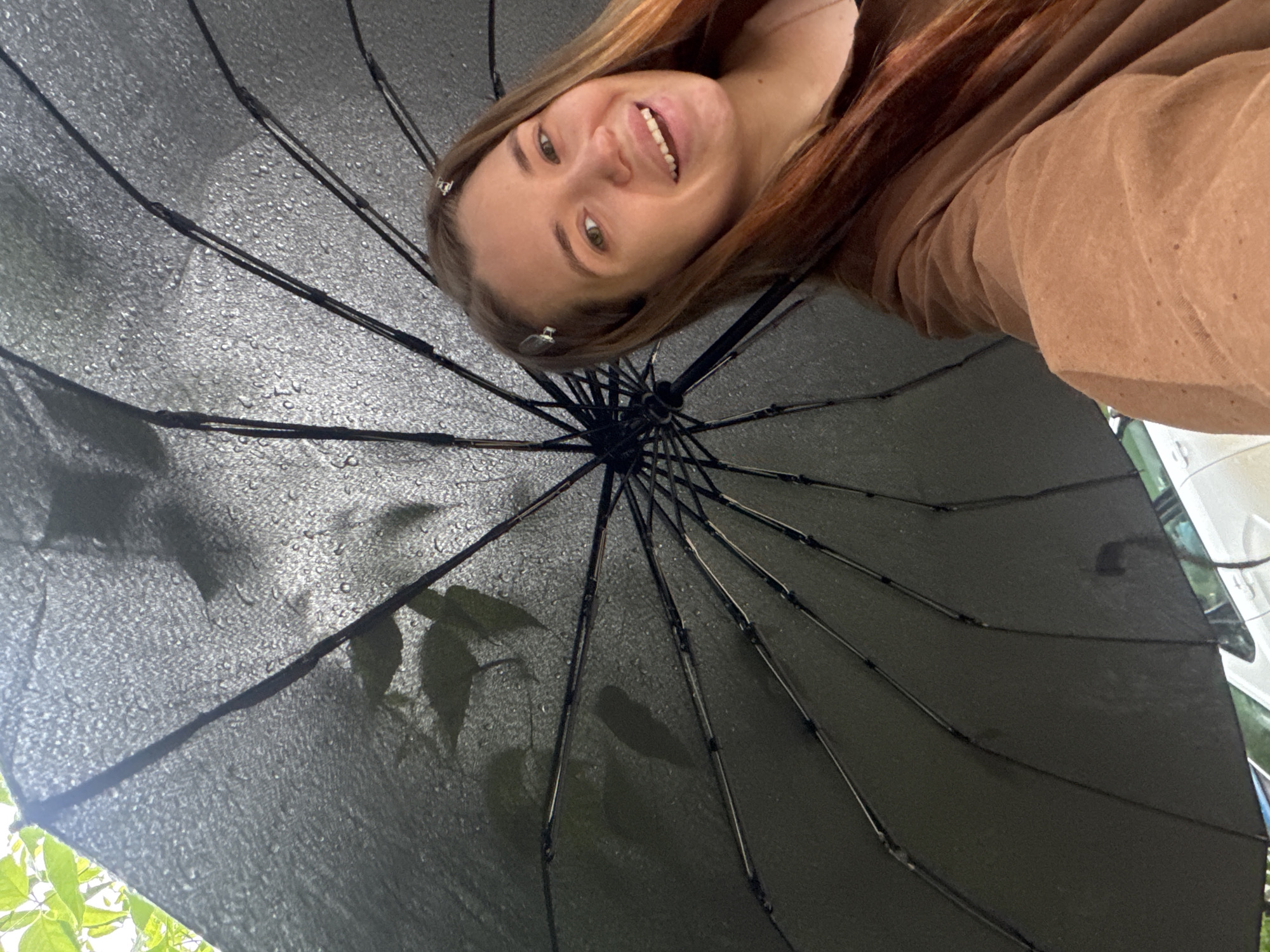 A person with reddish-brown hair and light skin smiles at the camera while standing under a black umbrella. Water droplets cover the umbrella's surface. Shadows of leaves and tree branches are visible through the umbrella. The person wears a tan or brown shirt or sweater. Parts of a vehicle and what appear to be leaves on trees are visible in the background. The location is most likely Chaikovsky, Russia.