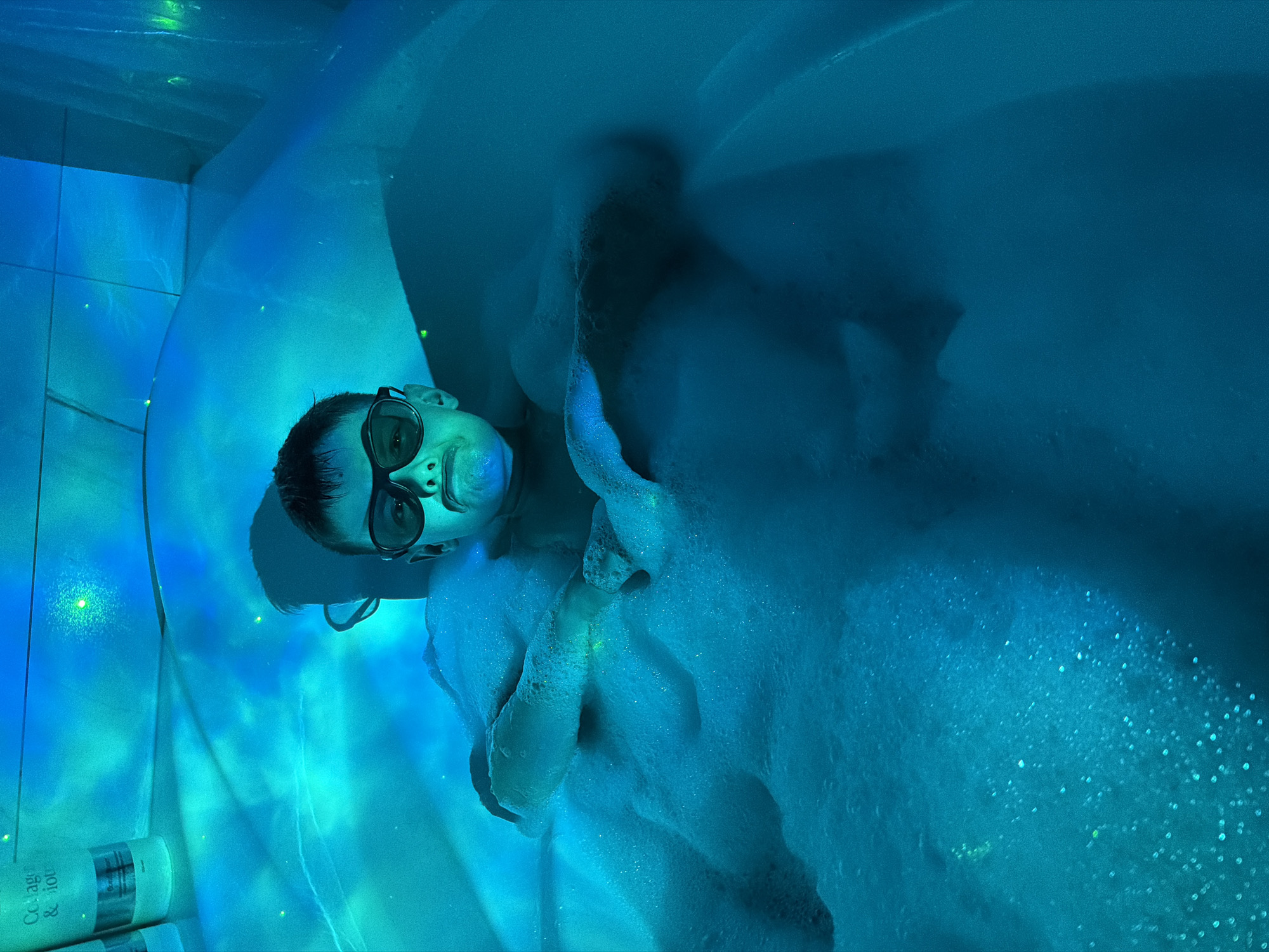 A person wearing glasses is submerged in a bubble-filled bathtub, possibly in Votkinsk, Russia. The environment has a bluish tint, giving a surreal appearance to the scene. A bottle of 'Collagen & Iou' can be seen to the left.