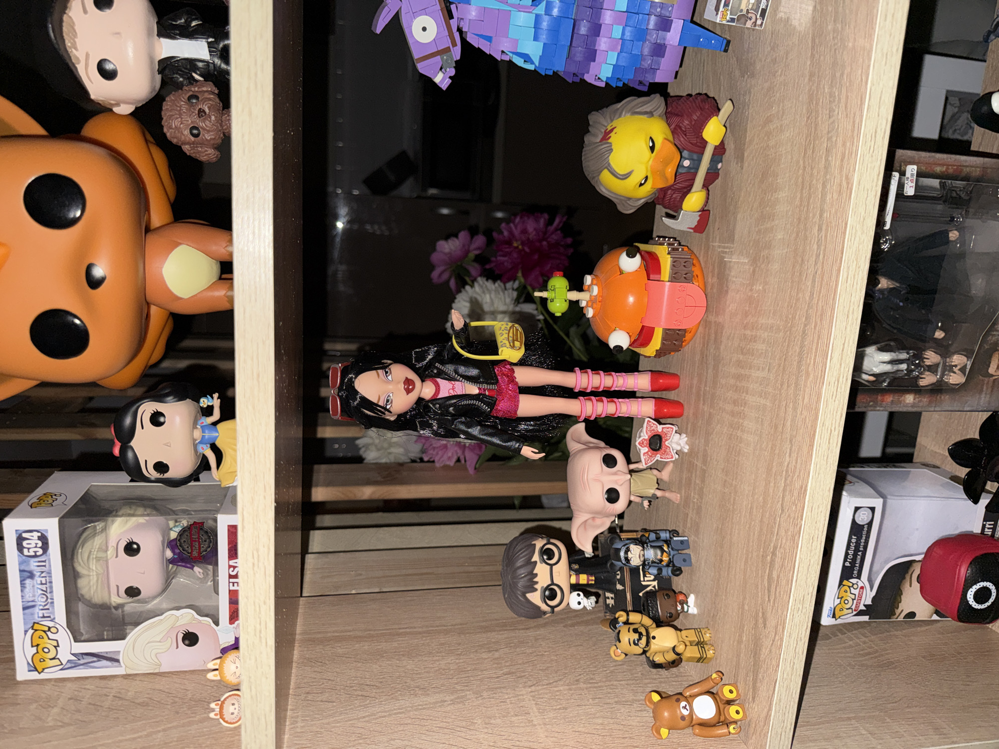 The image shows a collection of figurines and toys displayed on wooden shelves. On the top shelf, there are Funko Pop figures of Elsa and Snow White from Disney, as well as a large orange Funko Pop figure of what appears to be a Pokémon, and a small brown dog figurine.
The middle shelf features a variety of figures. On the left, there is a Harry Potter Funko Pop figure, a Rilakkuma figure, a Freddy Fazbear figure, and another smaller figure with a blue helmet. Further to the right, there is a Bratz doll in a pink outfit, a figurine of Dobby from Harry Potter, a Demogorgon figure from Stranger Things, a Burger King Fortnite figure, a duck-like figure holding an axe, and a purple llama figure made of LEGO bricks. Behind the Bratz doll, there are some flowers.
The bottom shelf displays more items, including a Funko Pop box labeled "Producer," a red Squid Game figure, and other figures partially visible in boxes.
The overall environment appears to be an indoor display, possibly a shelf in a room or a store. The lighting is somewhat dim, with some reflections visible on the surfaces. The image does not contain any information that would allow for the determination of its location in the city of Khimki, Russia.