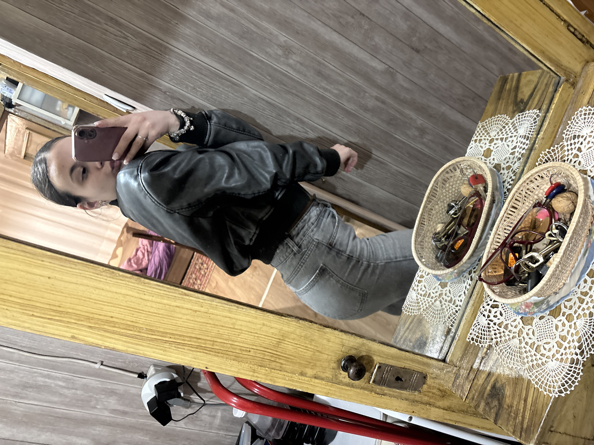 A woman is taking a mirror selfie. She is wearing a black leather jacket and gray jeans. She is holding a rose gold-colored smartphone to take the picture. The mirror reflects a room with wood paneling. There is a wooden dresser or cabinet with two woven baskets containing keys, walnuts, and sunglasses. The dresser has two lace doilies under the baskets. A power strip with plugs and cords is visible near the bottom of the frame. The location is possibly in Radomir, Bulgaria.