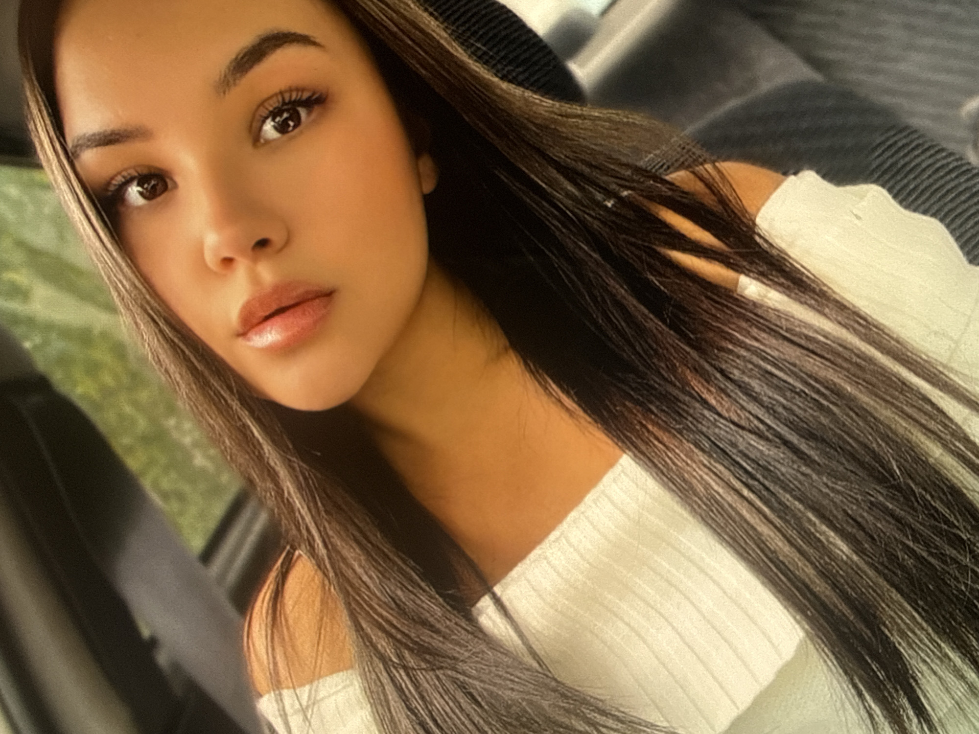 A young woman with long, dark brown hair, light brown skin, and light brown eyes is photographed at a close angle. She is wearing a white, off-the-shoulder, ribbed shirt. The background shows a car interior; specifically, a black car seat and a car window. The location is unknown, therefore I cannot provide details of Sofia, Bulgaria.
