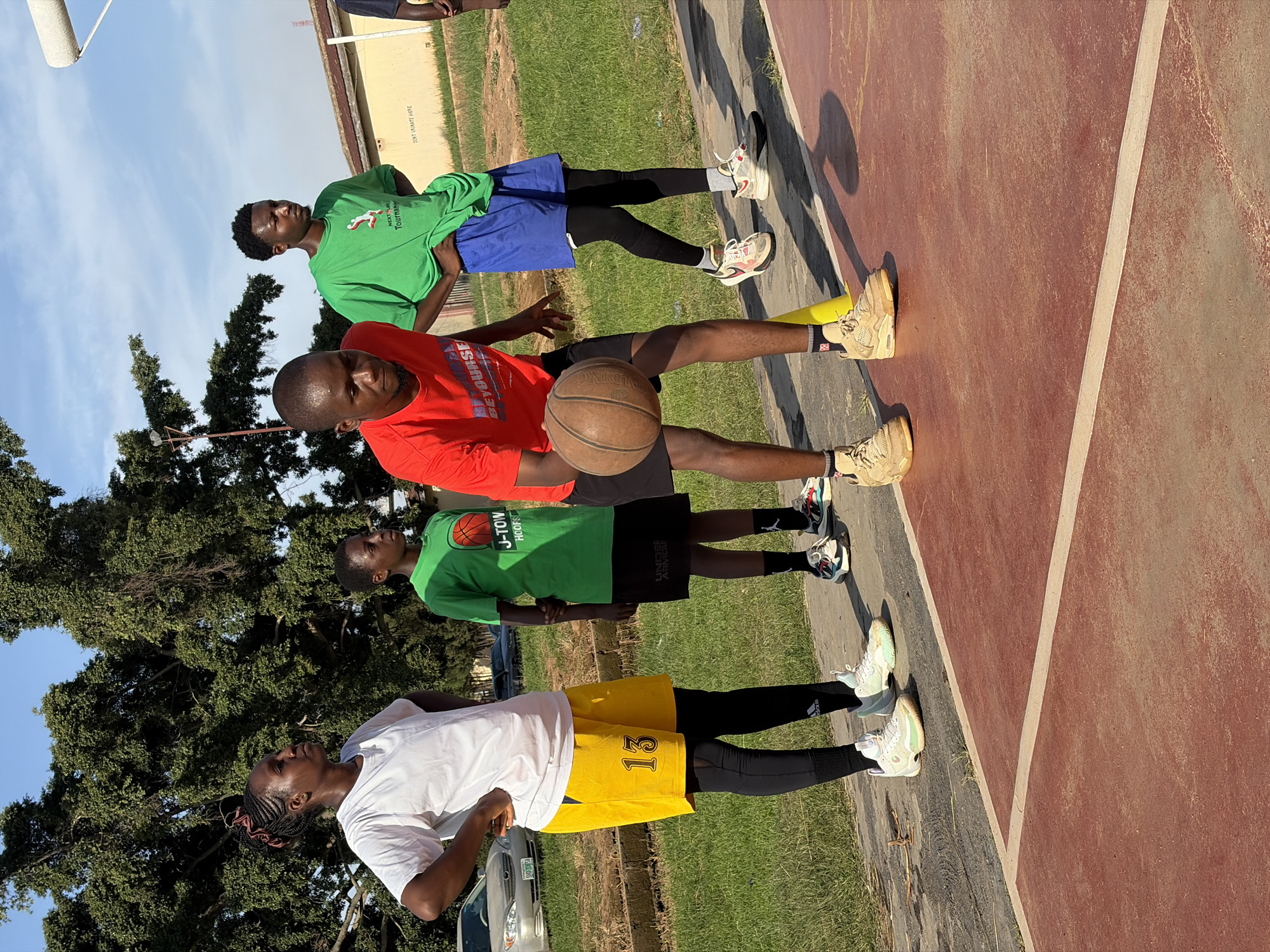 Four young people are practicing basketball on an outdoor court. The court appears to be a public facility with a red-colored surface and white lines. In the background, there are trees, grass, a building, and parked cars. The sky is clear and blue.
On the left, a young woman wearing a white t-shirt and yellow shorts with the number "13" on them is standing with her hands on her hips. She is wearing black leggings and white athletic shoes.
Next to her, a younger individual in a green t-shirt and black shorts is also standing.
In the center, a young man in a red t-shirt is dribbling a basketball. He is wearing black shorts and tan athletic shoes. A yellow traffic cone is near him.
To the right, another young man in a green t-shirt and blue shorts is standing with his hands on his hips. He is wearing black leggings and athletic shoes.
The overall impression is of a casual basketball practice session in an urban environment, possibly in Jos, Nigeria, given the context. The lighting suggests it is daytime, likely late afternoon due to the long shadows.