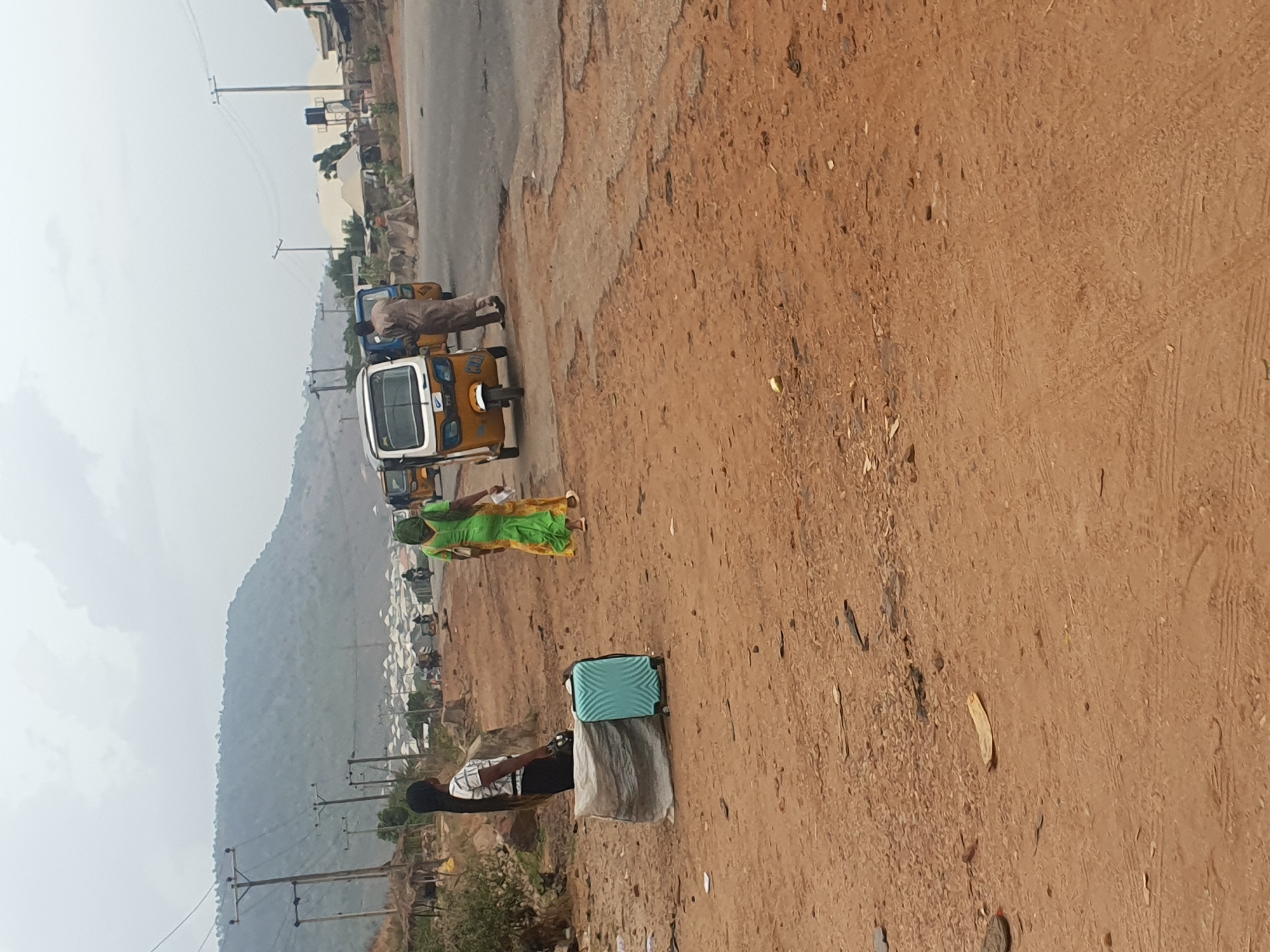 The image captures an outdoor scene along a road, typical of a semi-urban or rural area, consistent with Lokoja, Nigeria. In the foreground, a wide, reddish-brown dirt path or shoulder occupies the right side, textured with tire tracks and small debris. To its left, a narrower, darker paved road runs parallel.
Two individuals are visible on the dirt path. Closer to the viewer, a person wearing a checkered shirt and dark trousers stands next to a light blue, hard-shell suitcase. Further down the path, another person in a vibrant green and yellow traditional garment is walking or standing.
On the paved road, a line of three-wheeled vehicles, commonly known as keke napep or tuk-tuks, are present. One prominent yellow and white keke is partially visible, with a person in light brown attire standing near it. Other vehicles are faintly seen in the distance.
The background is dominated by a hazy, large mountain range under an overcast sky, suggesting a cloudy day but clear daylight. Below the mountain, some residential structures or a small settlement are discernible. Utility poles with power lines are scattered throughout the mid-ground. The scene portrays daily life and transportation activities, with people and vehicles coexisting in an environment that blends natural landscape with developing infrastructure. No specific text is clearly legible in the image.