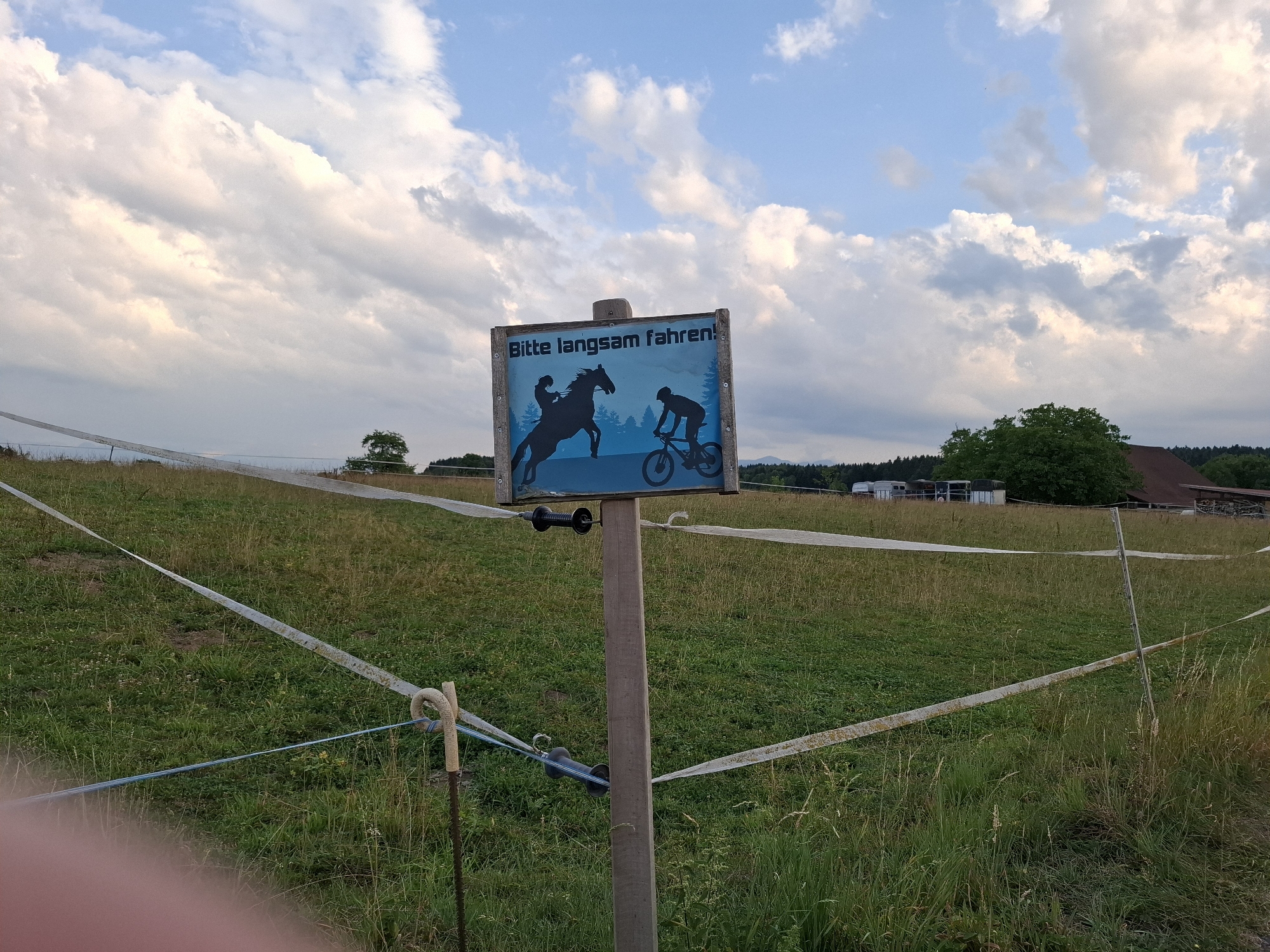 The image captures a rustic wooden signpost in a grassy field under a cloudy sky. The sign, made of wood and featuring a blue background, displays a silhouette of a person riding a horse on the left and a silhouette of a person on a mountain bike on the right, with trees in the background. The text on the sign reads "Bitte langsam fahren!", which translates to "Please drive slowly!". The field is bordered by a white electric fence, with a blue wire running parallel to it. In the distance, a building with a brown roof and several trailers are visible. The overall setting suggests a rural or agricultural area, possibly a park or a trail. The photograph appears to have been taken in the city of Hochdorf, Switzerland, given the contextual information.