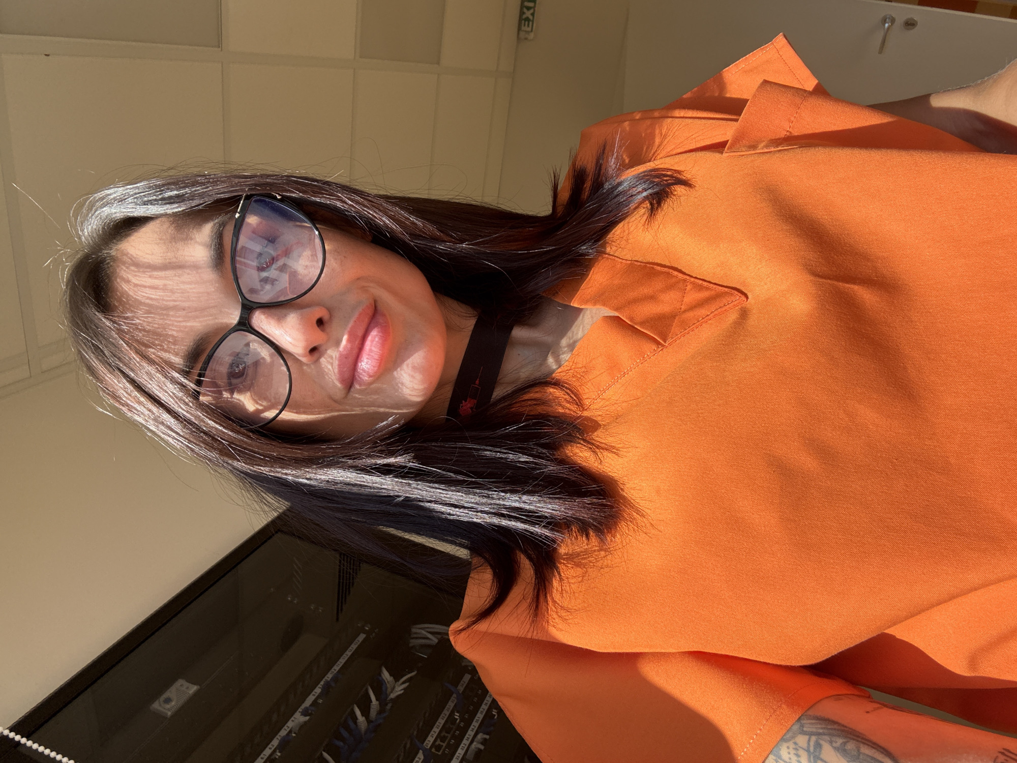 A selfie shows a woman wearing an orange scrub top and black-rimmed glasses. Her hair is dark brown with some lighter streaks and is falling over her shoulders. A black choker with a red pendant is visible around her neck. In the background, a server rack is visible, along with a ceiling and a door with a silver handle. An "EXIT" sign is also visible on the wall. The lighting suggests it is daytime, with sunlight casting shadows. The location is identified as Pleven, Bulgaria.