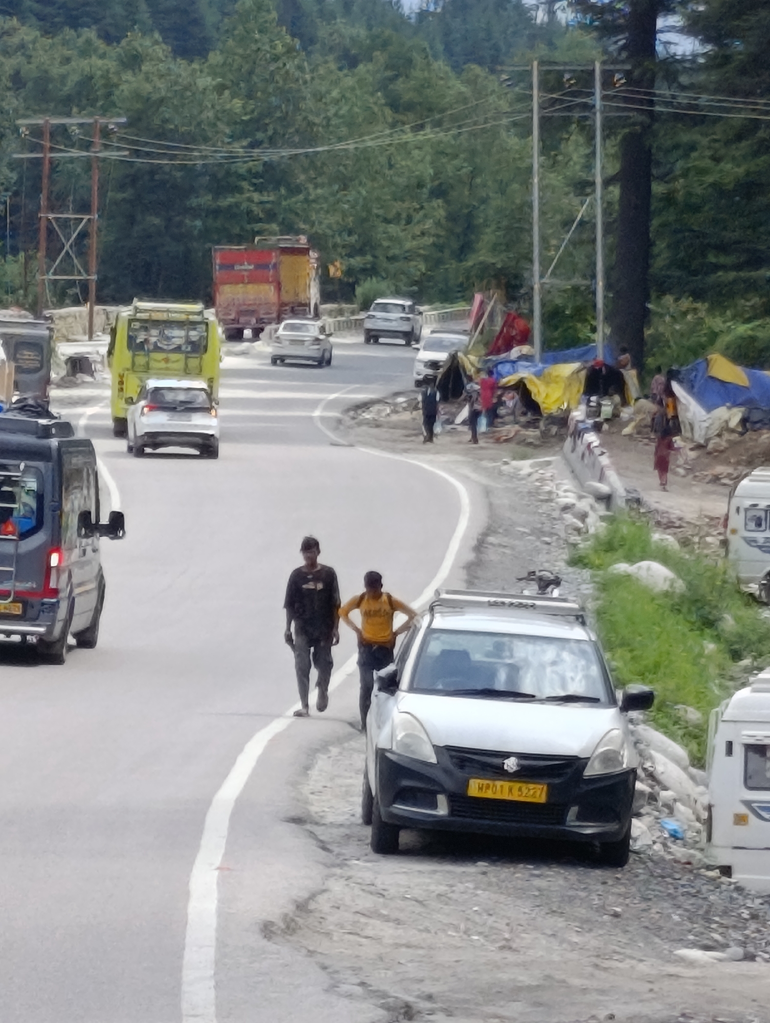 This image captures a street scene in Manali, India, likely near a busy road passing through a forested area. Several vehicles are present, including a blue van on the left, a yellow bus, a white car, and a white van further down the road. A silver car with a yellow license plate (MP01 K 5227) is parked on the right side of the road. Two men are walking on the road in the foreground, one wearing a dark t-shirt and the other a yellow t-shirt. In the background, a few tents or temporary shelters are visible with people around them, suggesting a roadside market or settlement. The environment is characterized by a paved road, a rougher shoulder with rocks and vegetation, and a backdrop of dense green trees and hills. Power lines are strung across the scene, supported by utility poles.