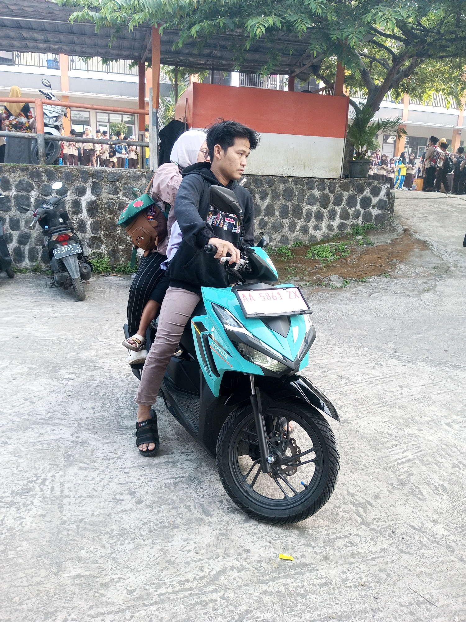 A young man is riding a turquoise Honda scooter, with two young children as passengers. The man, wearing a black hoodie and light brown pants, is looking to the side. The front passenger, a young child wearing a green beret and a brown backpack, is also looking to the side. A second young child, wearing a white hijab and a pink headscarf, is in front of the man, also looking to the side. They are on a paved area in front of a building with a stone wall. In the background, a group of schoolchildren in uniform are standing near a fence. The environment appears to be outdoors in a school or community setting.