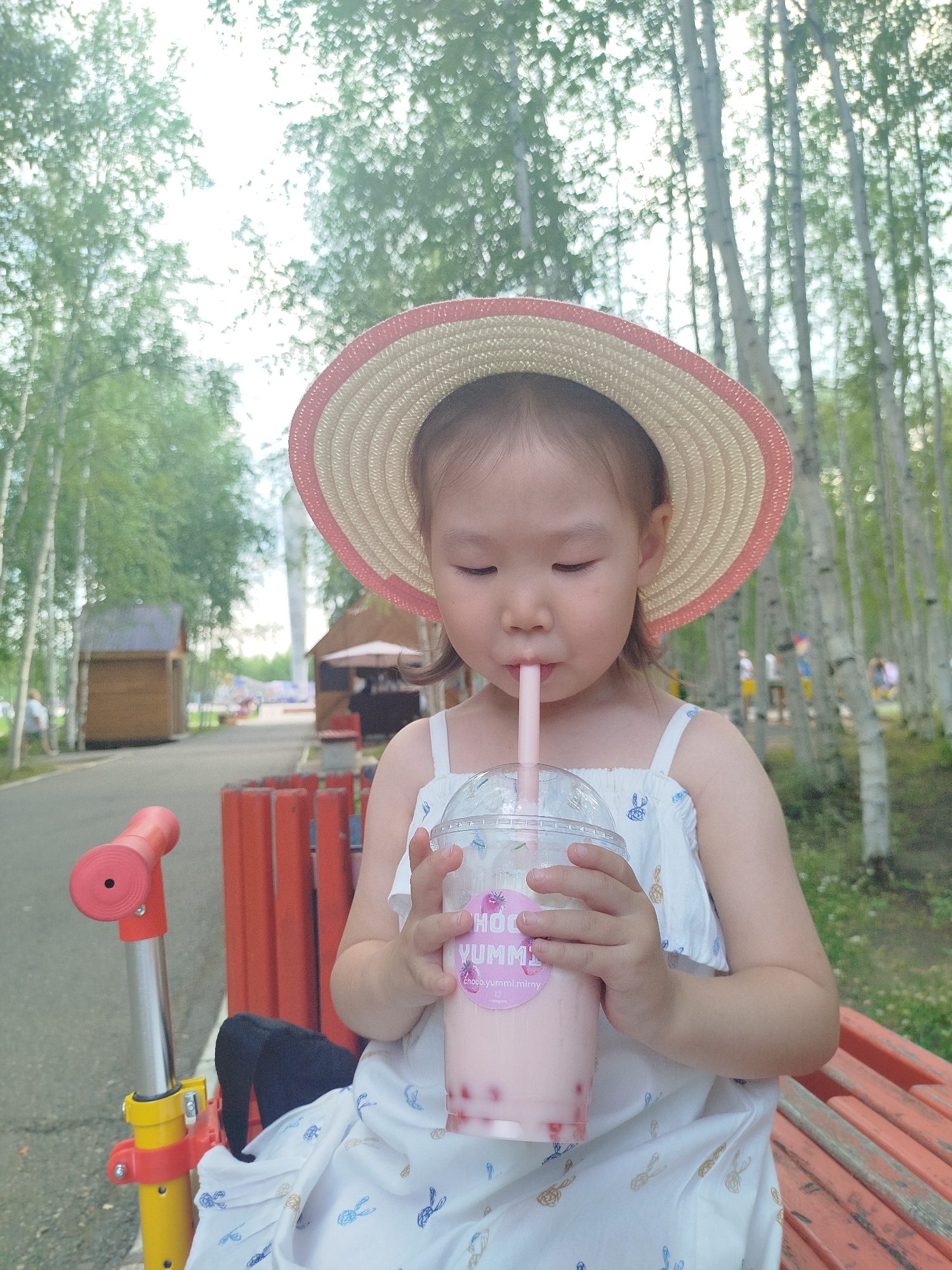 A young Asian child wearing a straw hat and a white dress patterned with blue and brown rabbits is seated outdoors and drinking a pink beverage from a plastic cup with a straw. The child is holding the cup with both hands, and the straw is extended to their mouth. In the background, there is a blur of trees, with some wooden structures and a red fence visible. To the left of the child, a red and yellow scooter is partially visible. The setting appears to be a park or a recreational area in Mirny, Russia, given the presence of birch trees commonly found in the region and the implied context of a public space. The lighting suggests it is daytime.