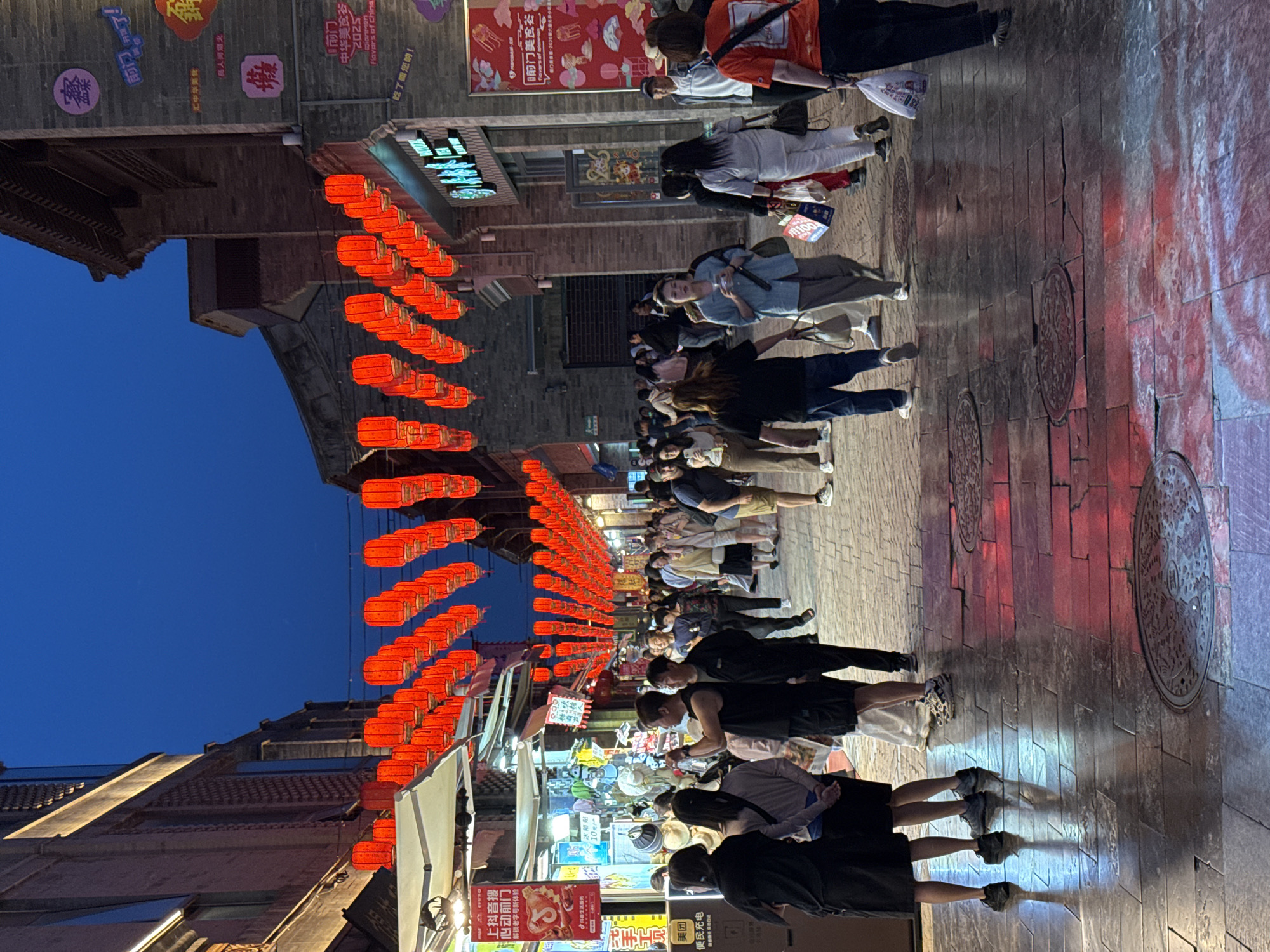 The image captures a lively night scene in what appears to be a commercial street in Dongcheng District, China. The street is lined with traditional-style buildings, adorned with rows of vibrant red lanterns that create a festive ambiance. A variety of people are walking along the street, many of them wearing face masks. Shops are visible, some with signs in Chinese characters. The ground appears to be paved with brick or stone. Manhole covers are visible in the road. The sky above is dark blue, indicating it is either late evening or night. The light from the street reflects on the ground.