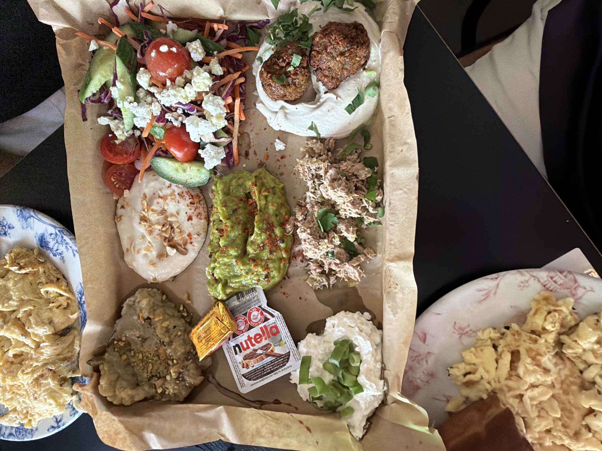 A high-angle shot shows a brown parchment paper-lined tray filled with various foods. The tray includes a salad with cabbage, carrots, cucumbers, tomatoes, and feta cheese; a white creamy spread topped with chopped nuts; guacamole sprinkled with red pepper flakes; tuna salad garnished with parsley; a dollop of white cream cheese with sliced green onions; and two falafel balls atop a creamy spread. A small packet of Nutella is also visible in the tray. To the left, a blue and white floral-patterned plate holds a portion of what appears to be pasta. To the right, a pink and white floral-patterned plate contains scrambled eggs. The background includes a dark surface, possibly a table, and a glimpse of a light-colored cushion. Given the style of food and ingredients, this is possibly a meal in a restaurant or cafe somewhere in the city of Tel-Aviv, Israel.