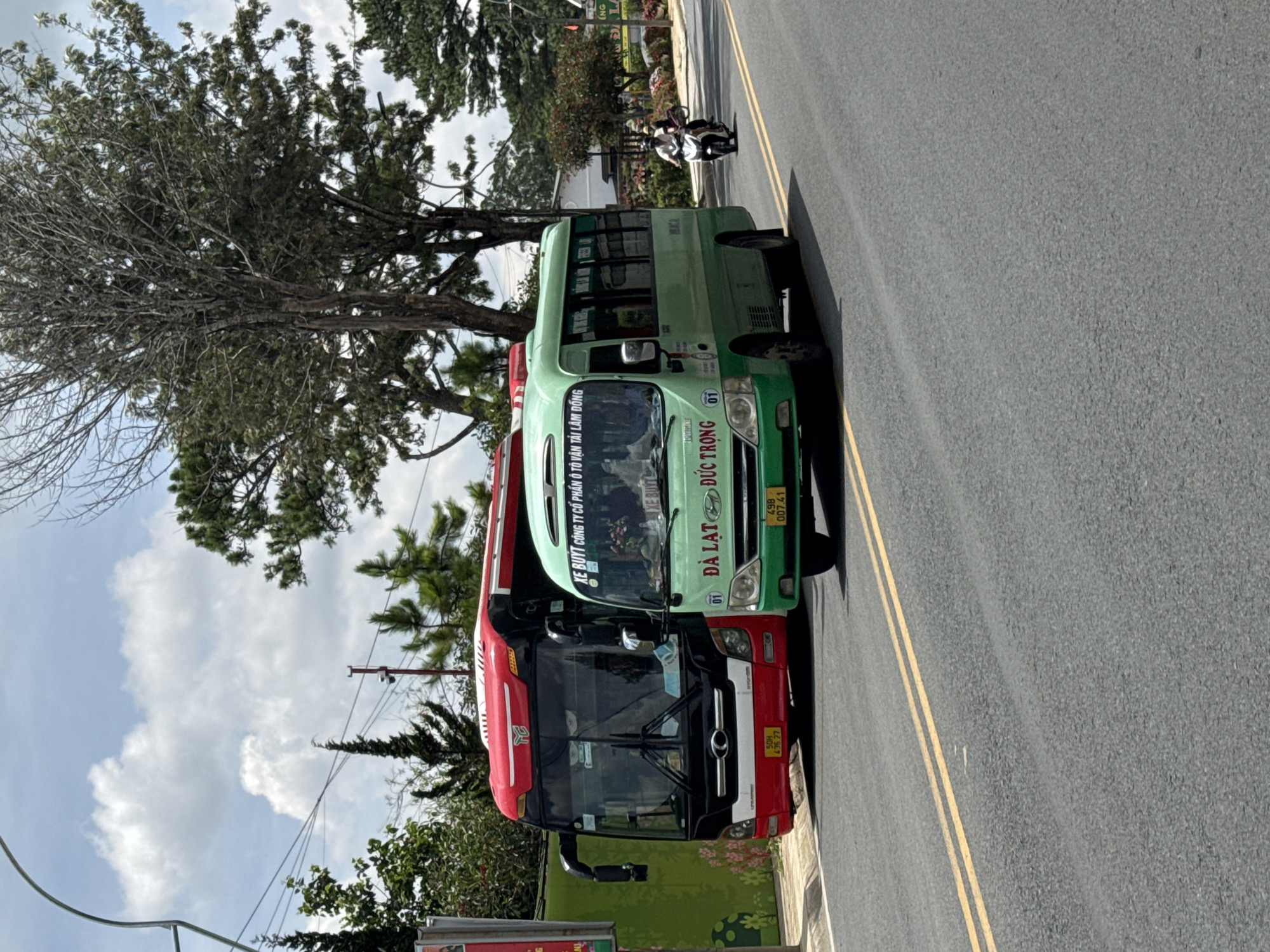 The image shows two buses parked on the side of a paved road with double yellow lines. The bus in the foreground is red and white, and the bus behind it is green. Both buses have visible text. The red bus has "50H 436.27" on its license plate. The green bus has "XE BUÝT CÔNG TY CỔ PHẦN Ô TÔ VẬN TẢI LÂM ĐỒNG" written on its side, along with "ĐÀ LẠT ĐỨC TRỌNG" and "49B 007.41" on its license plate.
The scene is outdoors, with trees and a cloudy sky visible. In the background, there appears to be a person riding a motorcycle, and some buildings. The lighting suggests it is daytime, possibly late morning or early afternoon. The overall impression is a typical roadside scene in Vietnam, likely in or around the city of Da Lat, given the text on the buses. There are no strong emotional cues evident.