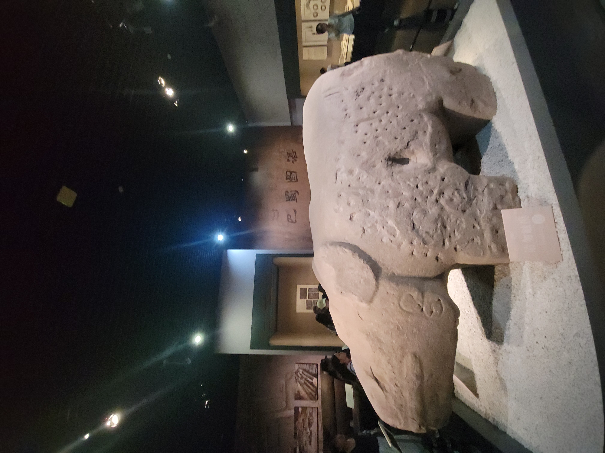 This is an indoor photograph of a stone sculpture of an animal, possibly an elephant, displayed in a museum setting. The sculpture is a light brown, weathered stone and appears to be quite large. It is resting on a base covered with small, white gravel. The sculpture is positioned in the foreground, filling a significant portion of the frame.
In the background, on the left side, there are framed pictures or artifacts mounted on the wall, and a few visitors are partially visible. To the right of the sculpture, there is a wall with some writing in Chinese characters. Further to the right, display cases with more artifacts and exhibits are visible, along with a few more people browsing the museum. The ceiling of the museum is dark and features several spotlights illuminating the exhibits.
The overall environment suggests a cultural or historical museum. The presence of the sculpture, the displays, and the visitors indicate it is a public exhibition space.
The sculpture is located in the Qingyang District of China, as confirmed by the inscription on the small sign in front of the sculpture, which reads "庆阳." This suggests the sculpture is an artifact from or related to the Qingyang region of China.