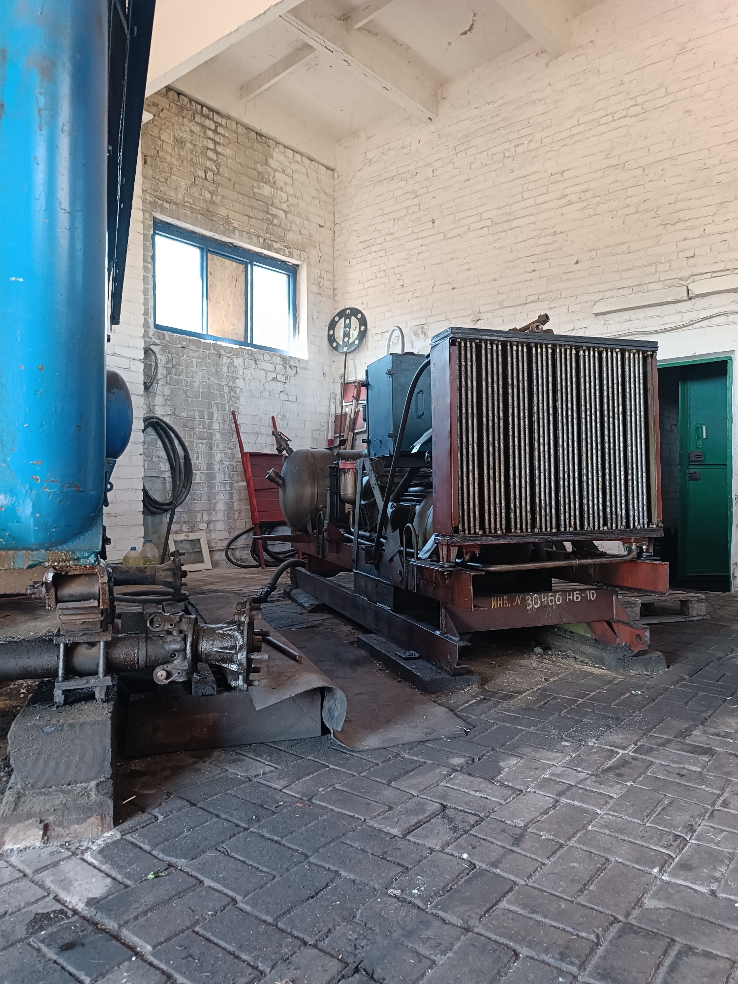 In Vuhlehirsk, Ukraine, inside a sparsely lit industrial building with white brick walls and a concrete floor, a large, rusted metal radiator unit sits mounted on a red metal base. The radiator is situated in the center of the frame, with its vertical metal fins dominating its front surface. To the left of the radiator, a large blue pipe extends upwards and out of frame, and to its right, a closed green door is visible. In front of the radiator, on its red base, is a stencil with Cyrillic text and numbers that read "ИНВ. № 30466 НБ-10". The overall environment appears to be a workshop or storage area, with various mechanical components and equipment visible in the background. The lighting is dim and casts long shadows, suggesting either early morning or late afternoon.