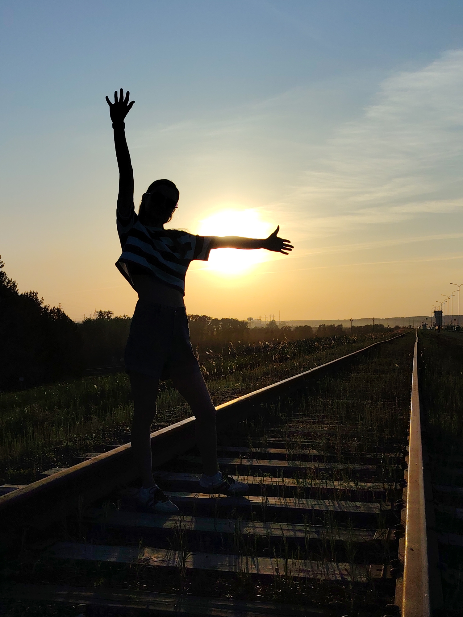 A silhouette of a woman stands on a railway track against a sunset. She has one arm raised straight up and the other stretched out to the side. She is wearing a striped top and shorts. The tracks stretch into the distance, flanked by vegetation. The setting sun is visible between the railway tracks. The scene is Chaikovsky, Russia.
