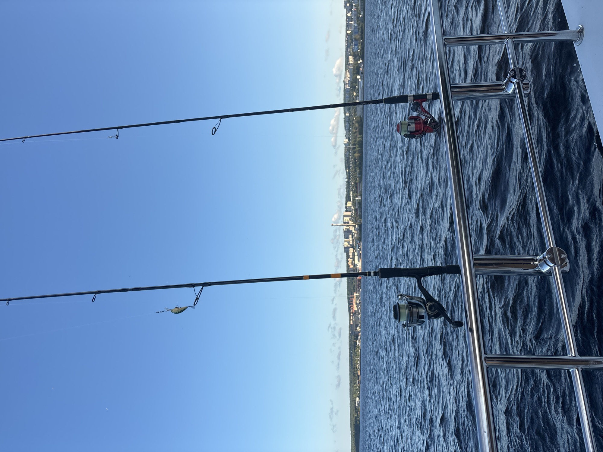 Two fishing rods are planted in holders on the deck of a boat, with the water of a large lake and a distant city skyline visible beyond. The fishing rods are tall and slender, made of black material with orange accents on one. The left rod has a lure attached to its line, which dangles in the air. The reels on the rods are metallic, one silver and the other red. The water is a deep blue, with small waves creating a textured surface. In the background, a city with numerous buildings, some appearing to be industrial structures with smokestacks, is spread along the treeline of the far shore. The sky above is a clear, bright blue. The scene is likely set on Lake Onega, overlooking the city of Petrozavodsk, Russia.