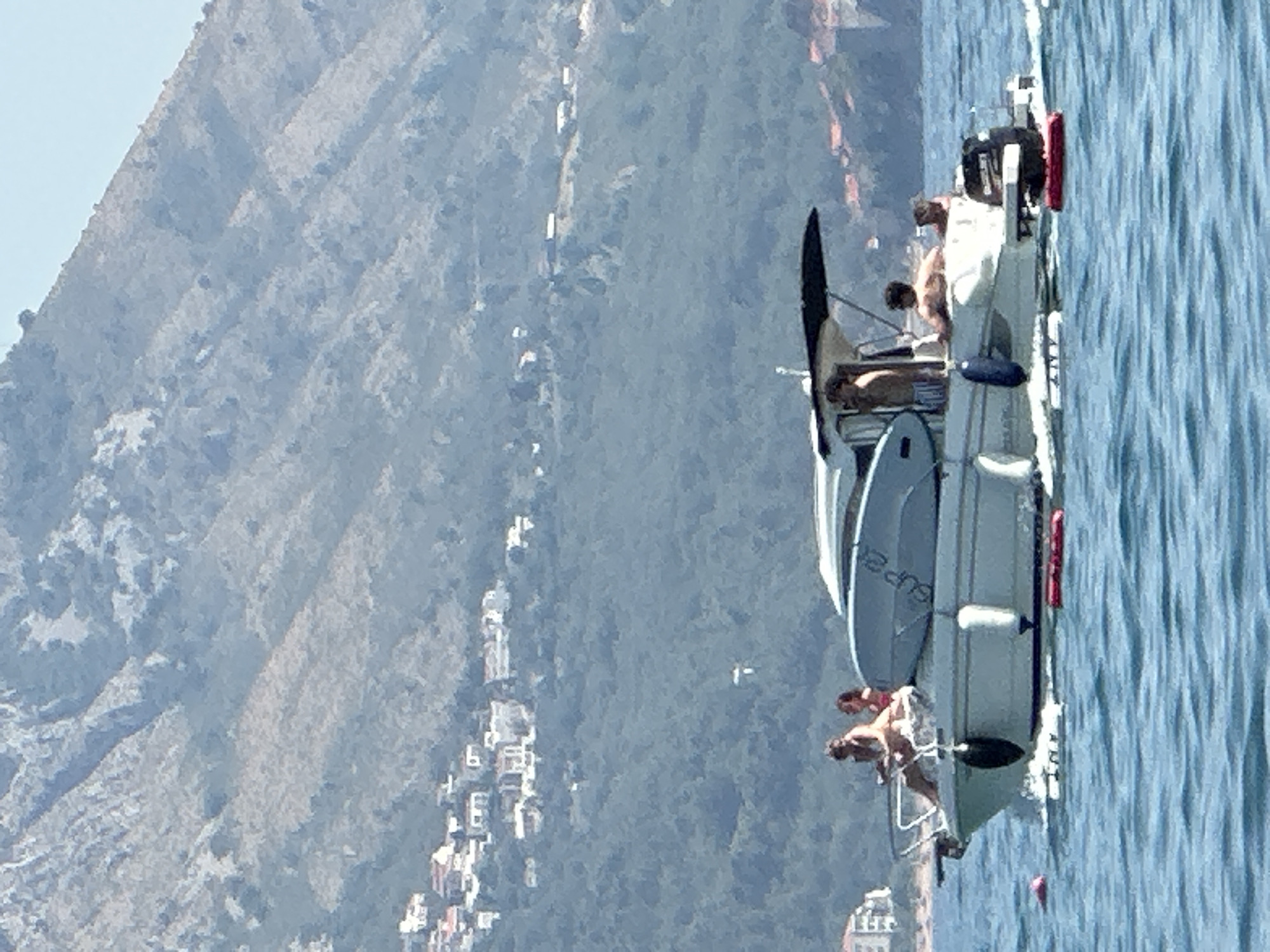 A small white motorboat with a dark canopy is sailing on blue water. There are at least five people on the boat. Two women, appearing to be sunbathing, are seated at the bow. A man is standing near the helm, and two other individuals are seated further back. A surfboard is attached to the side of the boat. In the background, a lush green hillside is dotted with white buildings, and a large, rocky mountain rises behind it. Red and pink buoys are visible in the water near the boat. The sky is clear and blue. The location is identified as Boreti, Montenegro.