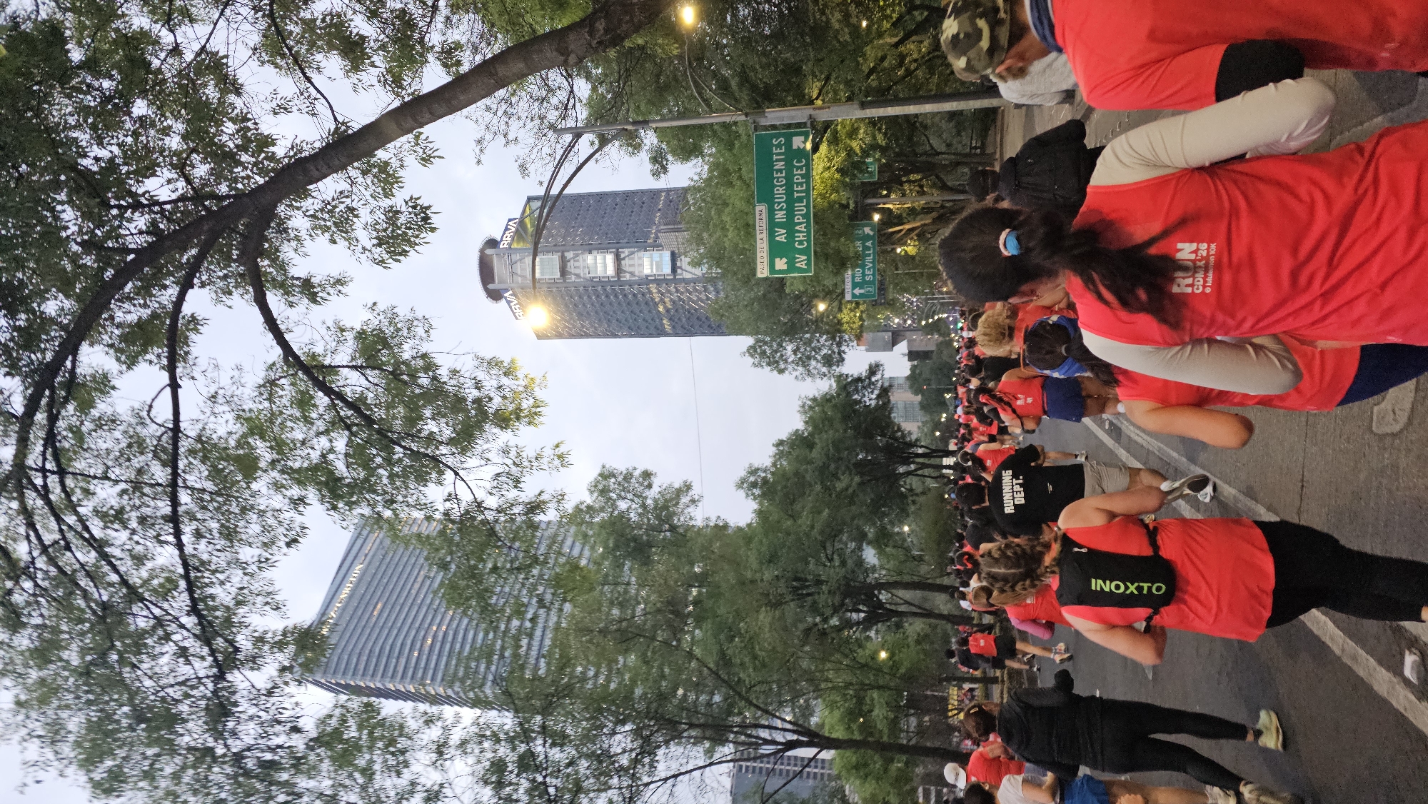 The image shows a large group of people participating in a running event in Mexico City, Mexico. Many participants are wearing red shirts and running along a street lined with trees. Buildings are visible in the background, including the BBVA Tower. Street signs indicate that the race route is along Paseo de la Reforma, with connections to Avenida Insurgentes, Avenida Chapultepec, and Rio Sevilla. The event may be associated with the "CDMX '26" and "lululemon 10K" based on the text on some of the participants' shirts.