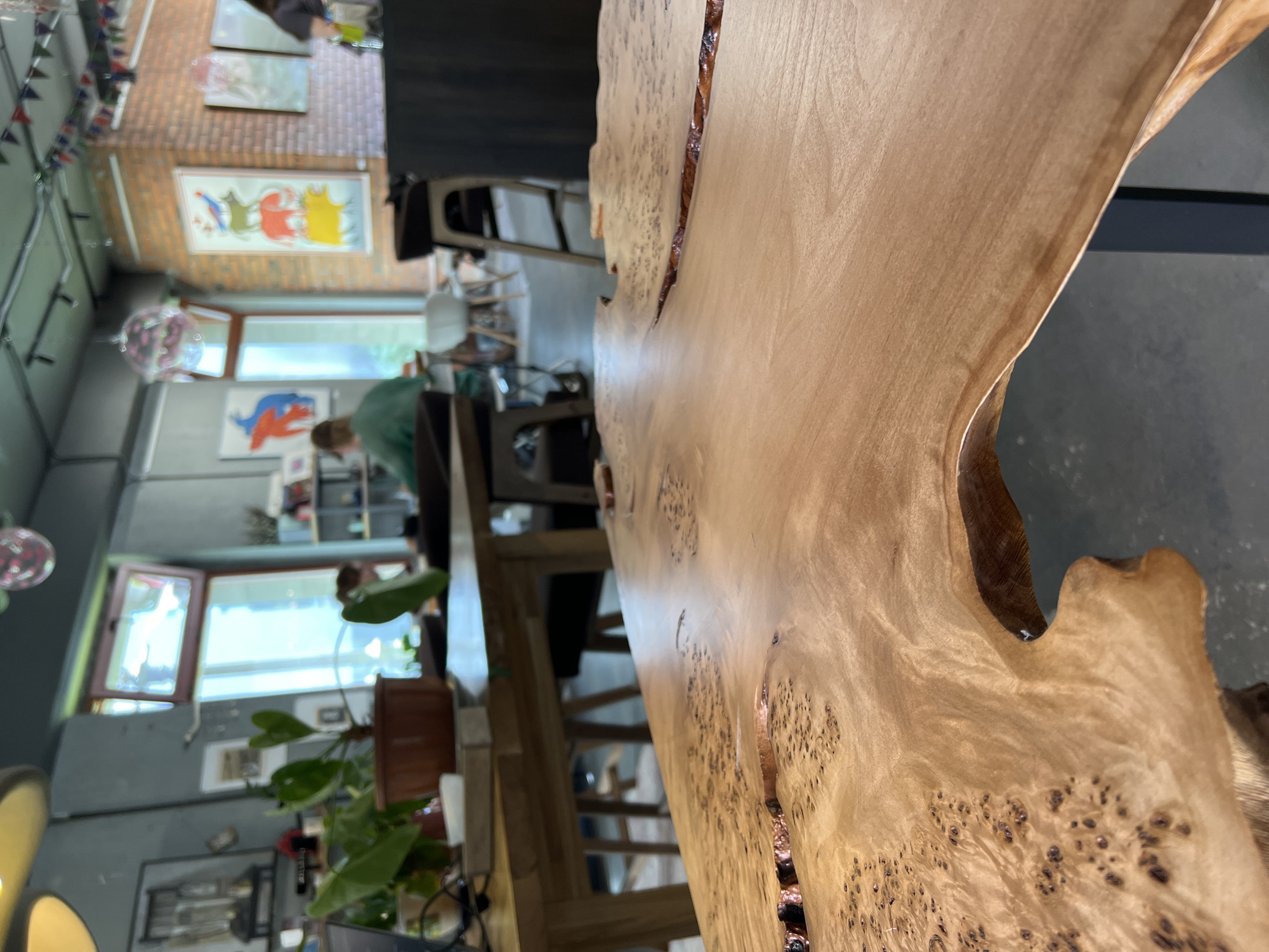 The image depicts the interior of a cafe. In the foreground, a light-colored, polished wooden table with a natural, uneven edge is prominently featured, showing prominent wood grain and burl patterns. The background is blurred but reveals a seating area with tables and chairs, and patrons. Some artwork is visible on the walls, including paintings. A plant is present on a table in the middle ground. The overall atmosphere suggests a modern, possibly artistic cafe setting. The image does not provide any explicit information to pinpoint a specific location in Saint Petersburg, Russia.