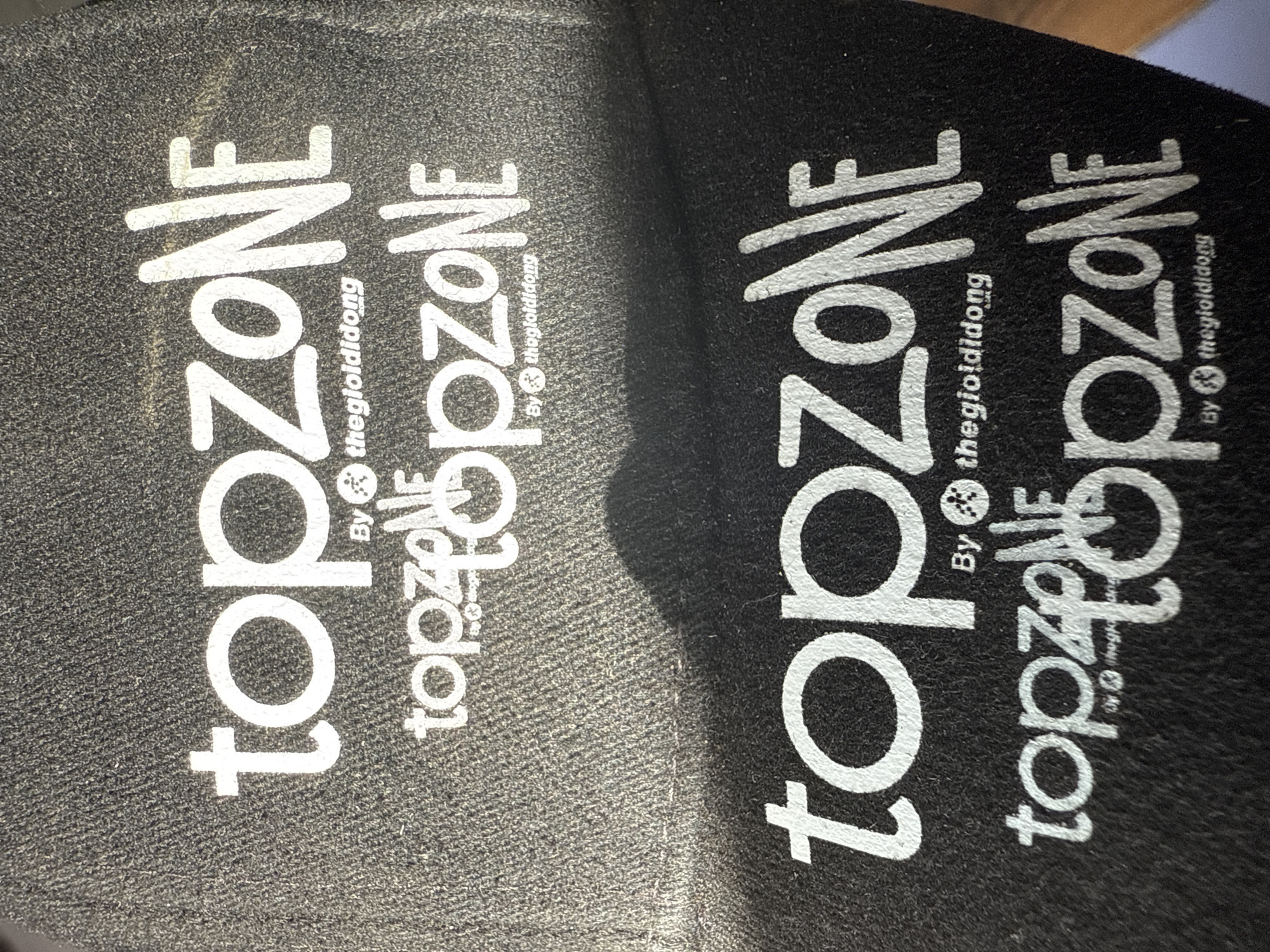 This image shows two black shoe inserts with the words "topZONE" printed in white and stacked vertically. Underneath "topZONE" is the phrase "topZONE" in a smaller font, also printed in white. Next to the smaller "topZONE," the words "By thegioididong" are printed in an even smaller font, accompanied by a small logo. The inserts are very similar and appear to be a pair. The material looks soft, potentially fabric or foam. The environment is not determinable from the provided image.