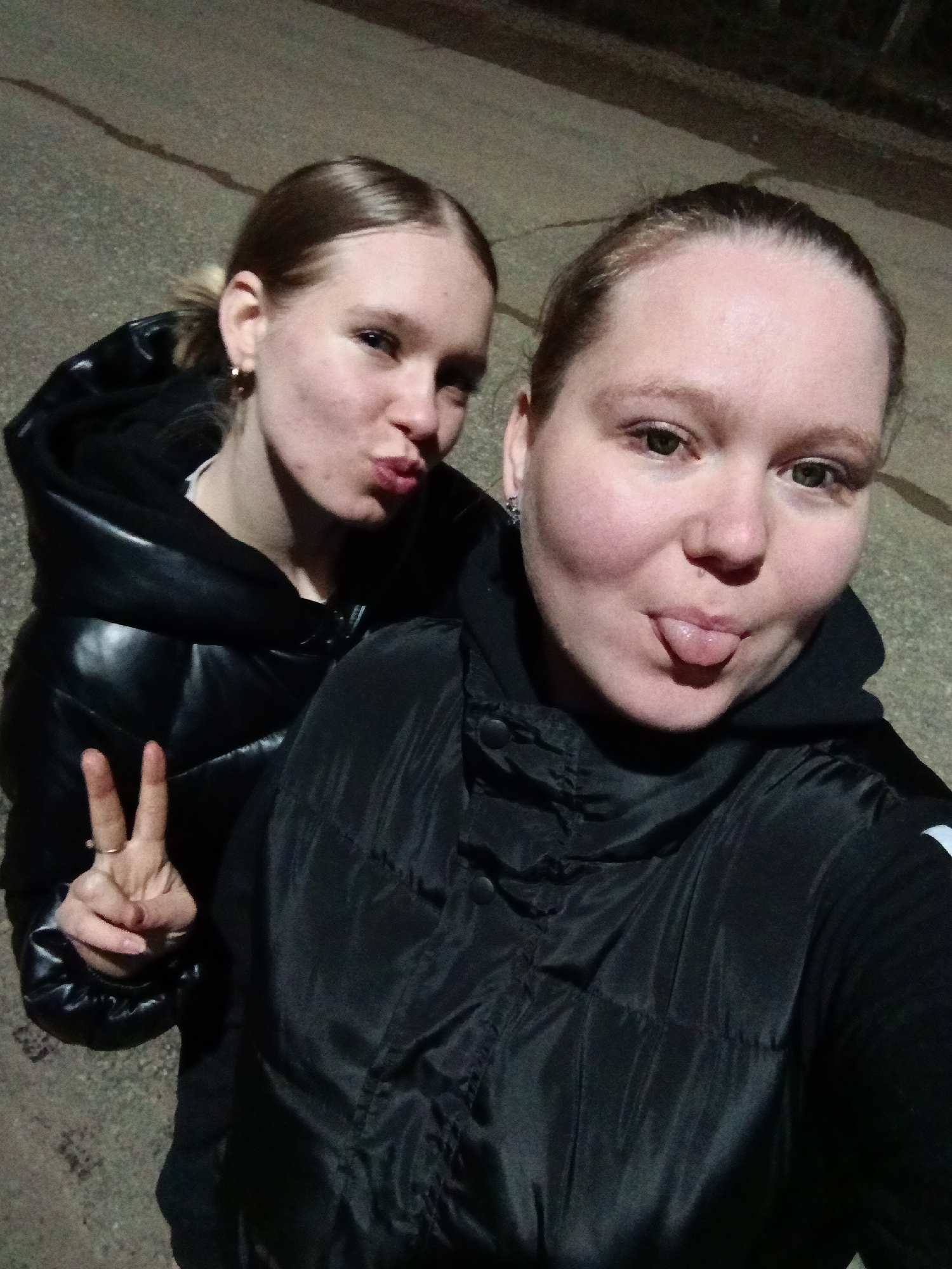 Two fair-skinned women pose outdoors at night on a paved surface. The woman on the left wears a shiny black puffer jacket and makes a peace sign with her left hand, puckering her lips. Her blonde hair is pulled back into a ponytail. She is wearing earrings and a ring. The woman on the right wears a black puffer jacket with a hood and sticks her tongue out. Her blonde hair is also pulled back. She wears earrings. The background is dark, and the location is inferred to be Rusalevka, Russia.