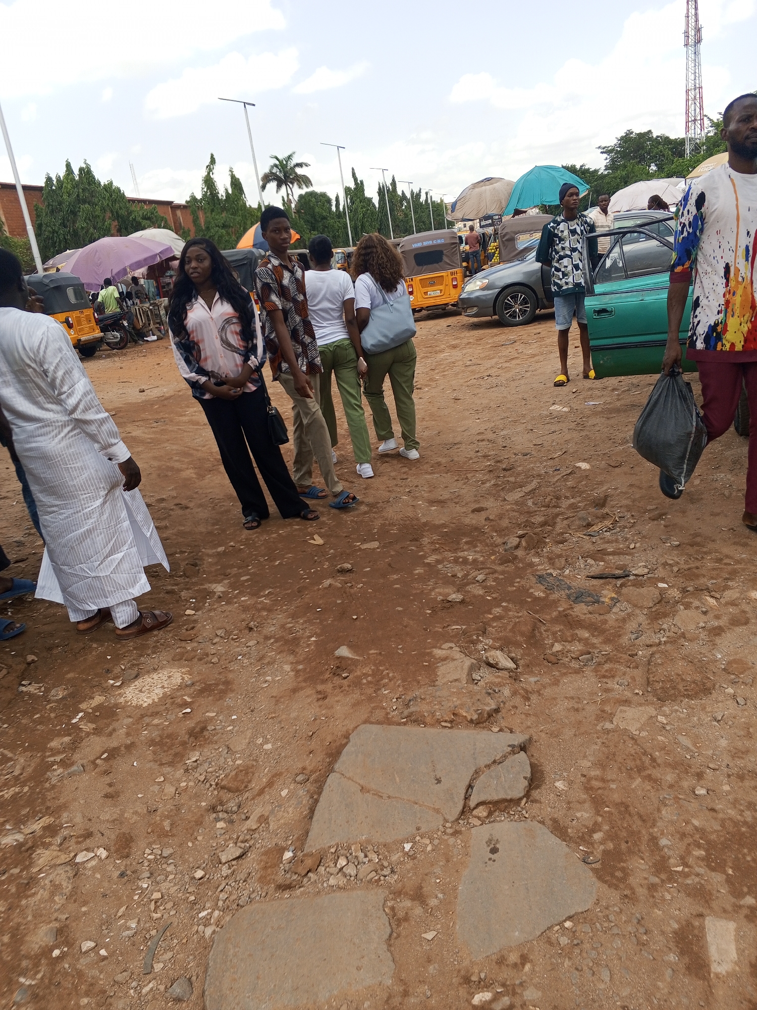 The image captures a dusty, unpaved area in Abuja, Nigeria, with several people present. In the foreground, the ground is composed of reddish-brown dirt interspersed with rocks and fragments of broken paving.
Several individuals are visible: a woman in a patterned blouse and black pants stands near a person in a patterned shirt, while behind them are two people with their backs turned. To the left, a person in white clothing is partially visible. Another person, dressed in a shirt with colorful patterns, is holding a black bag and walking towards the viewer.
Further in the background, there are parked cars, yellow tricycle taxis, and vendors beneath umbrellas. Streetlights and a telecommunications tower can be seen in the distance.