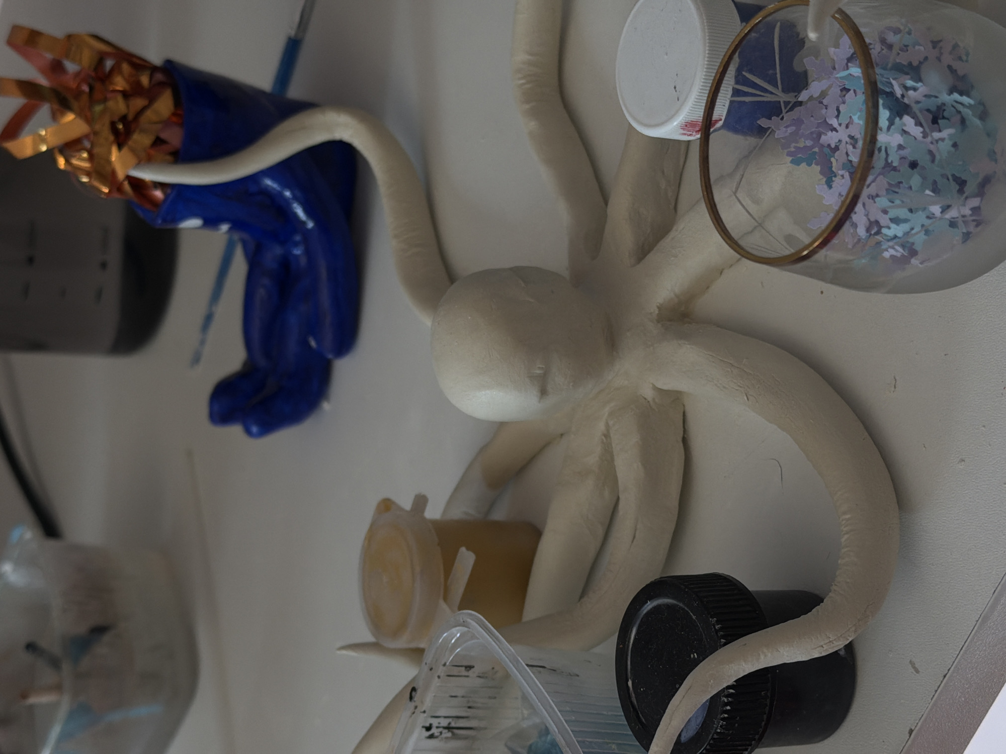 The image shows a close-up, overhead view of a white, clay octopus sculpture surrounded by various objects on a white surface. The octopus has eight tentacles, each interacting with different objects: one with a blue gloved hand holding gold strips, another with a white lid, a third with a clear glass container filled with purple and blue paper confetti, a fourth with a yellow jar, and a fifth with a black jar. A paintbrush lies near the blue gloved hand. Other partially visible containers and miscellaneous items are scattered around the octopus. The context hints at a creative or artistic workspace, potentially a studio. The image does not offer any information about a location in the city of Perm, Russia.
