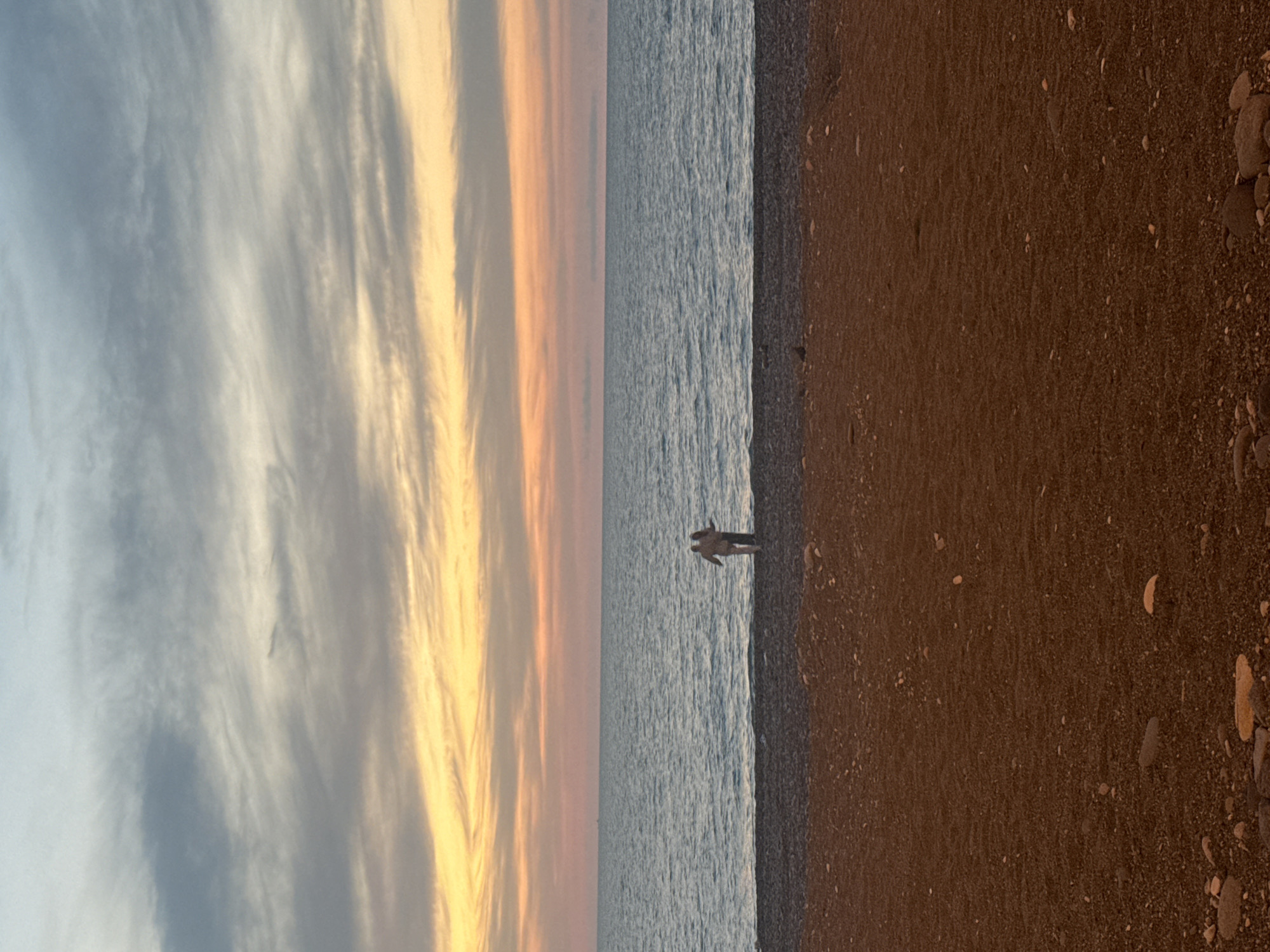 The image presents a landscape featuring a beach at sunset. From left to right, the sky is predominantly blue with streaks of white and orange clouds. Adjacent to the sky is the ocean, appearing calm with subtle ripples on the surface. A single person is standing at the edge of the water, their form silhouetted against the bright water. The beach is comprised of sand and pebbles. The image captures the natural elements and a lone individual in a serene, coastal setting. The location is likely Batumi, Georgia, as this city is known for its beaches and sunsets.