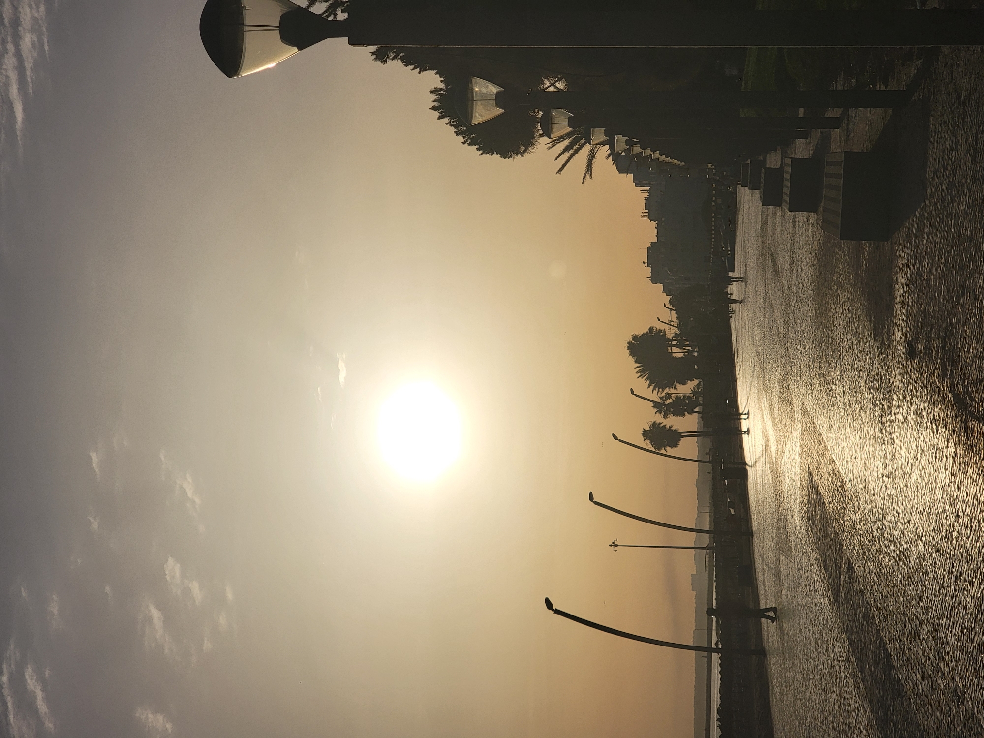 A long shot captures a promenade scene at dusk in Quarteira, Portugal. The sun, a bright orb in the hazy sky, casts a warm, golden glow across the paved walkway. The surface, made of small, irregular stones, reflects the light, creating a shimmering effect. The promenade is lined with lampposts, their dark silhouettes standing out against the bright sky. Some are modern and sleek, while others have a more traditional design. On the right side, a row of dark benches is visible, casting long shadows. Palm trees and other foliage can be seen behind the benches, adding a touch of greenery to the scene. In the distance, buildings of varying heights form a city skyline, softened by the dusky light. Several people are visible on the promenade, their figures silhouetted by the setting sun. One person is walking towards the viewer, while others are seen in groups further down the walkway, their movements indistinct in the low light. The sea or a large body of water is visible on the left side of the image, with a faint outline of structures on the horizon. The overall atmosphere is one of tranquility and end-of-day calm.