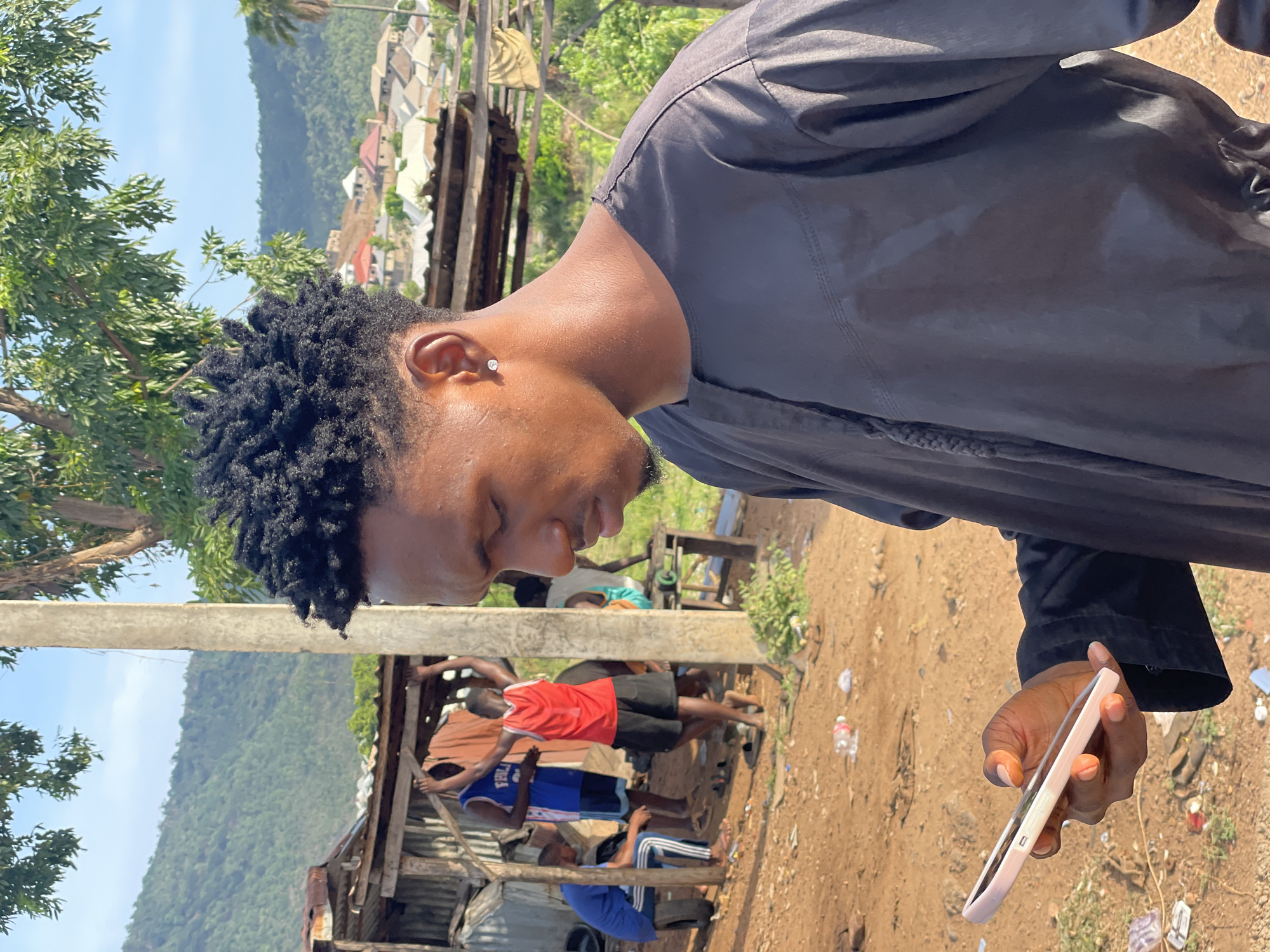 A man with dark, short, textured hair stands in an outdoor setting, holding a cell phone in his right hand. He is wearing a dark, long-sleeved shirt or jacket. The background reveals a somewhat cluttered scene with other individuals present. There are structures that appear to be makeshift or partially constructed, indicating a less developed urban environment. A dirt ground is visible, and a hillside or mountain is visible in the distance. The image is consistent with an outdoor location in Lokoja, Nigeria.