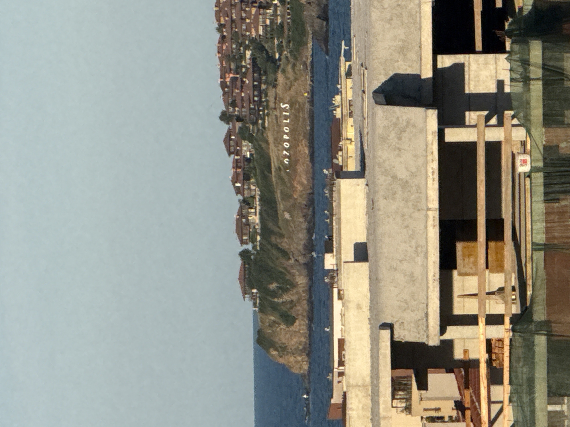 The image shows a coastal landscape with a cliff on the right side of the frame overlooking the sea. The cliff is covered in vegetation and dotted with a cluster of red-roofed buildings. A large white sign with the word "OZOPOLIS" is visible on the side of the cliff. In the foreground, there are several unfinished concrete structures, suggesting a construction site. The sky is clear and light blue. The image appears to be taken from an elevated position, looking towards the sea and the cliffside settlement. The overall scene depicts a combination of natural beauty and ongoing development in a coastal area.