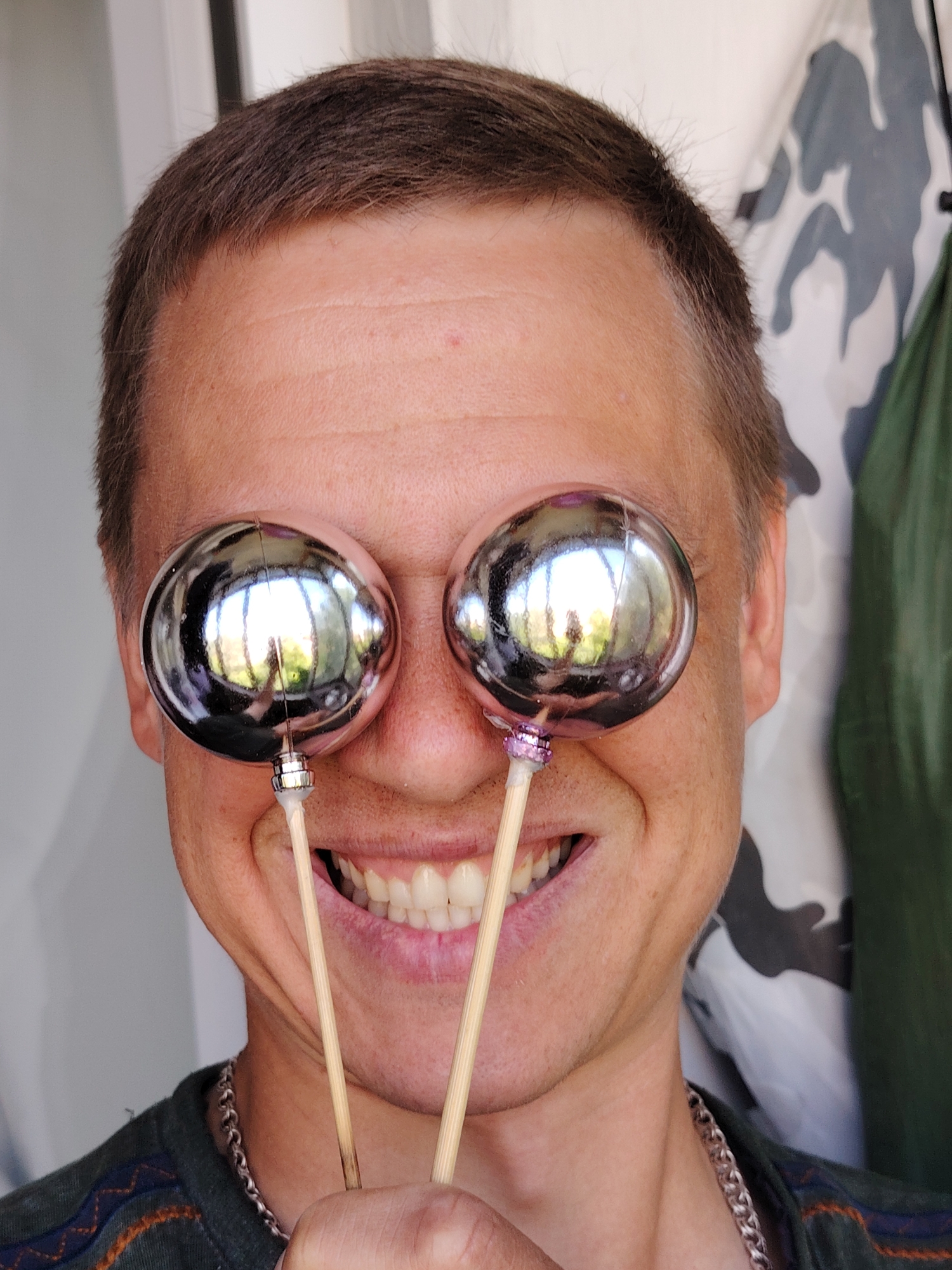 A man in Chaikovsky, Russia, has two shiny, spherical objects held up to his eyes. The objects are reflective, showing a distorted image of trees and sky. He is smiling broadly, revealing his teeth. The man is wearing a dark green shirt with a chain necklace. The background is out of focus, but appears to be an outdoor setting with some camouflage fabric visible.