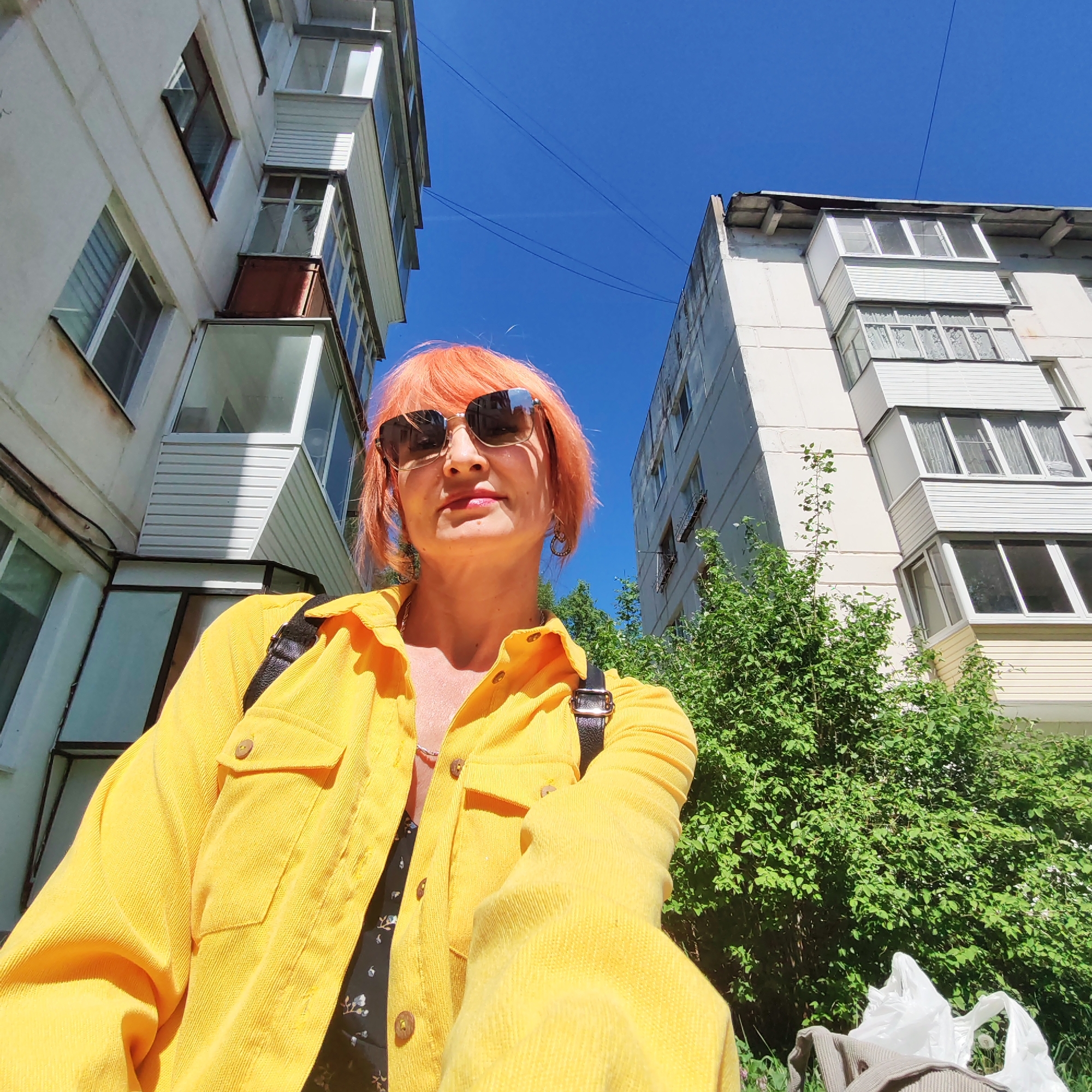 A woman with orange hair and sunglasses stands in the city of Chaikovsky, Russia, based on metadata. She wears a yellow corduroy jacket and a black backpack. Buildings are visible in the background, one with balconies. Green foliage is to her right. The sky is bright blue.