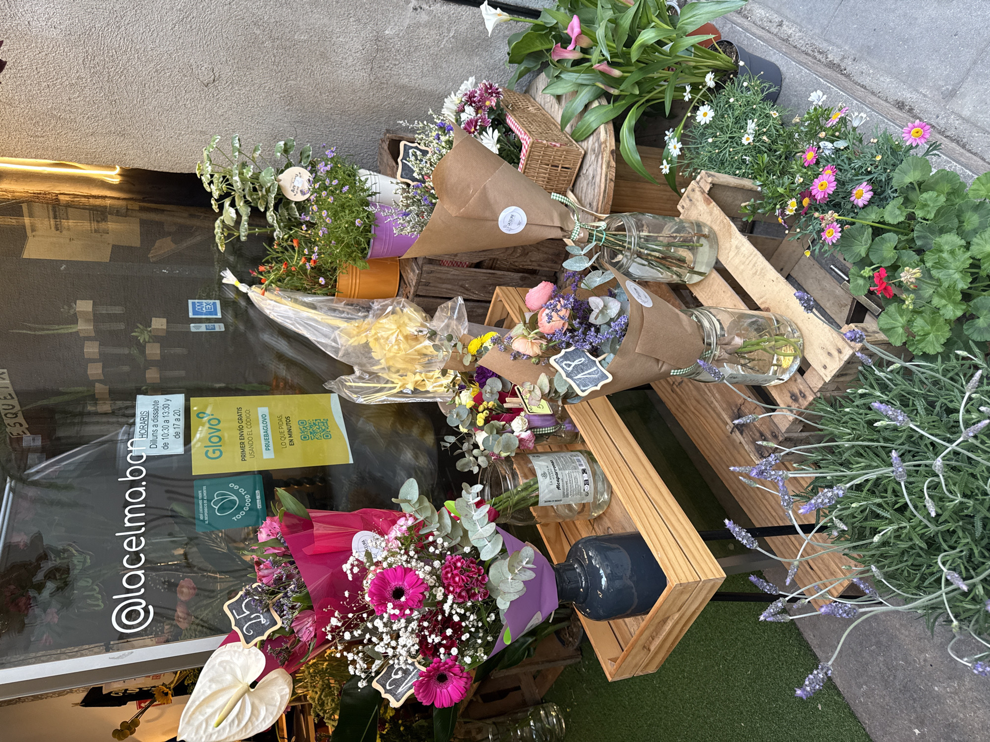 A street-level view shows a flower shop storefront, presumably in Barcelona, Spain, due to the Catalan language visible on the signage. The shop, "@lacelma.ba", displays an array of flower arrangements and potted plants on wooden crates and stands outside its glass doors.
The flower arrangements are diverse, some in brown paper wrapping with small chalkboard price tags, others in clear vases filled with water. One prominent bouquet features pink gerbera daisies and baby's breath, wrapped in pink and purple paper. Potted plants include white calla lilies, daisies, and lavender.
The storefront door has stickers indicating the shop's hours (Monday to Saturday), as well as partnership with the Glovo delivery service. The presence of a "Too Good To Go" sticker suggests the shop participates in food waste reduction initiatives. The overall scene conveys a small business with a focus on fresh flowers and sustainability.