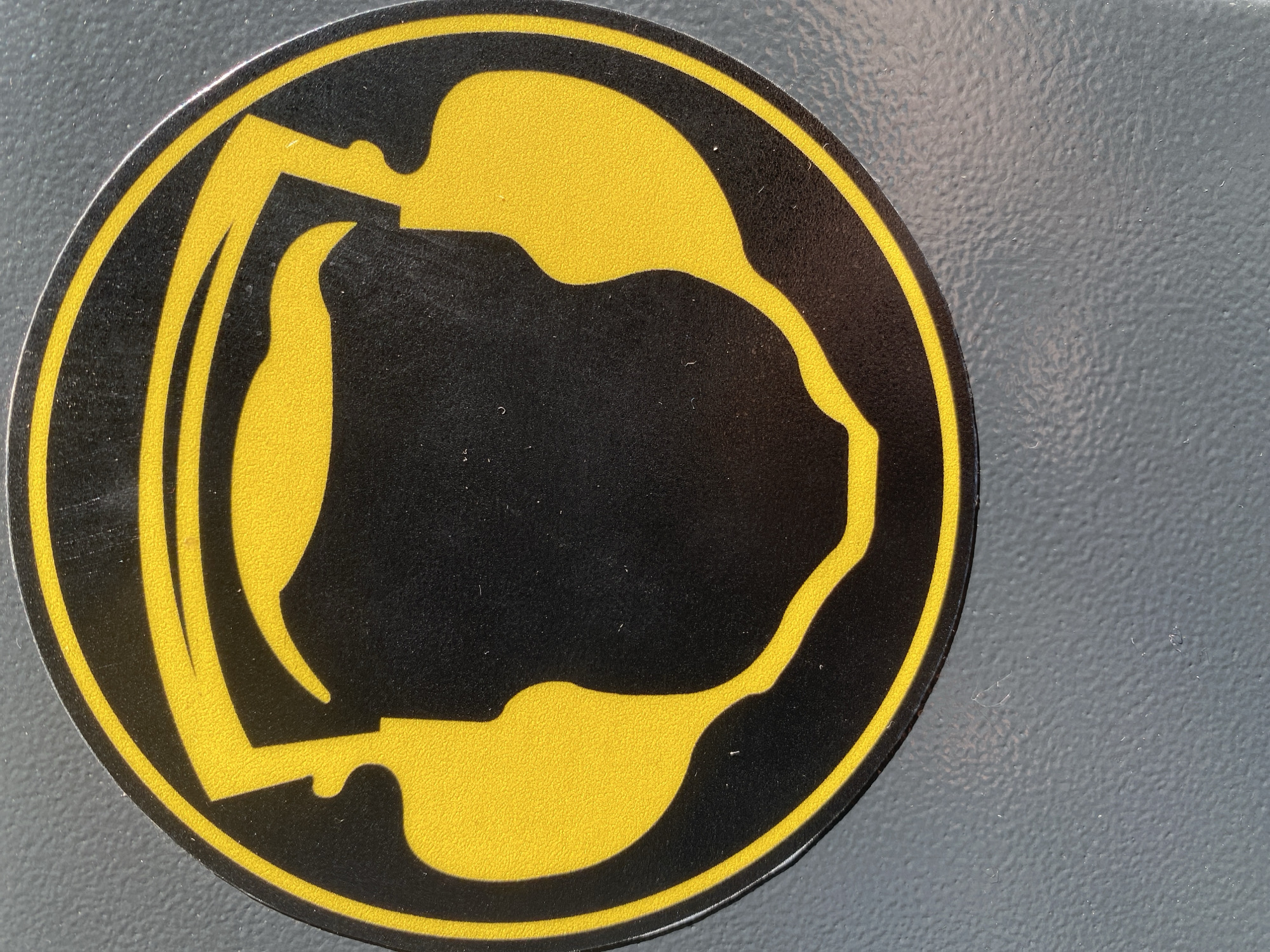 A circular sticker is prominently displayed, featuring a bold design in black and yellow. The sticker has a black interior with a thin, bright yellow border. Within the black area, abstract and organic yellow shapes dominate the design. On the left side, a stylized yellow figure with a distinct curved head and a flowing body occupies the space. This figure is connected to a larger, more amorphous yellow shape that fills the central and right portions of the sticker, characterized by rounded curves and an elongated section extending towards the right edge. The yellow areas exhibit a slightly pebbled or textured surface, suggesting the material of the sticker, and the black background also shows a subtle texture. The sticker is placed on a uniformly textured, matte surface, which appears to be gray or blue-gray. No human figures, specific objects beyond the sticker, or discernible activities are visible. The lighting is even, accentuating the textures. There is no visible text on the sticker.
