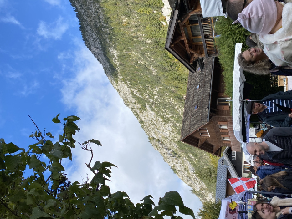 The image captures an outdoor market scene in Abondance, France, set against a backdrop of a prominent, steep mountain. The mountain's upper section features light grey rock faces, while its lower slopes are densely covered with dark green coniferous trees. The sky above is blue with scattered white clouds.
In the immediate foreground, dark green foliage partially obscures the upper left corner. Below the mountain, several traditional wooden chalet-style buildings are visible; a notable brown chalet with a gabled roof and windows is situated centrally.
The market area in the midground and foreground features multiple white canopy tents. One tent on the left displays a small Danish flag and a yellow sign indicating "CHAUSSETTES 15 €" (Socks 15 €). Numerous individuals populate the market. From left to right, a woman with light brown hair is partially visible. A bald man, wearing a dark vest over a grey long-sleeved shirt with an "HH" logo, stands facing slightly right, exhibiting a subtle smile. To his right, an older man in a dark flat cap, a blue and white striped shirt, and a dark jacket holds a dark umbrella or cane. Further right, a woman with curly blonde hair looks off to the right. In the extreme foreground on the right, a person's head, presumably an infant, is visible, wearing a light pink hooded garment with ear-like features. People are observed standing, walking, and interacting within the market environment.