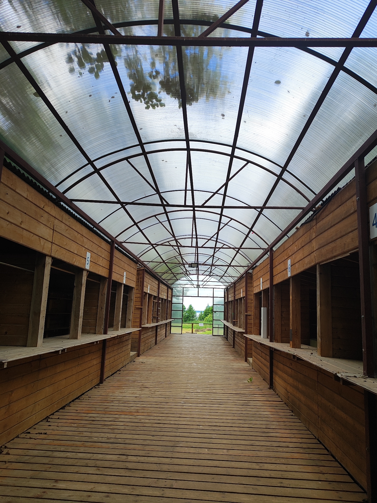 The image shows an outdoor market or pavilion in Никитская Слобода, Russia, with a long, covered walkway lined with wooden stalls. The walkway is constructed of wooden planks, and the stalls are made of wood, with open fronts. A translucent, arched roof made of corrugated polycarbonate supported by a dark metal frame covers the entire structure. The roof allows natural light to filter through, and reflections of trees and the sky are visible on its surface. Several stalls are marked with numbers, such as "5" and "4". The walkway leads to an open area at the far end, where greenery and a body of water are visible in the distance. The scene appears to be a public space, possibly for vendors to sell goods. There are no people visible in the image.