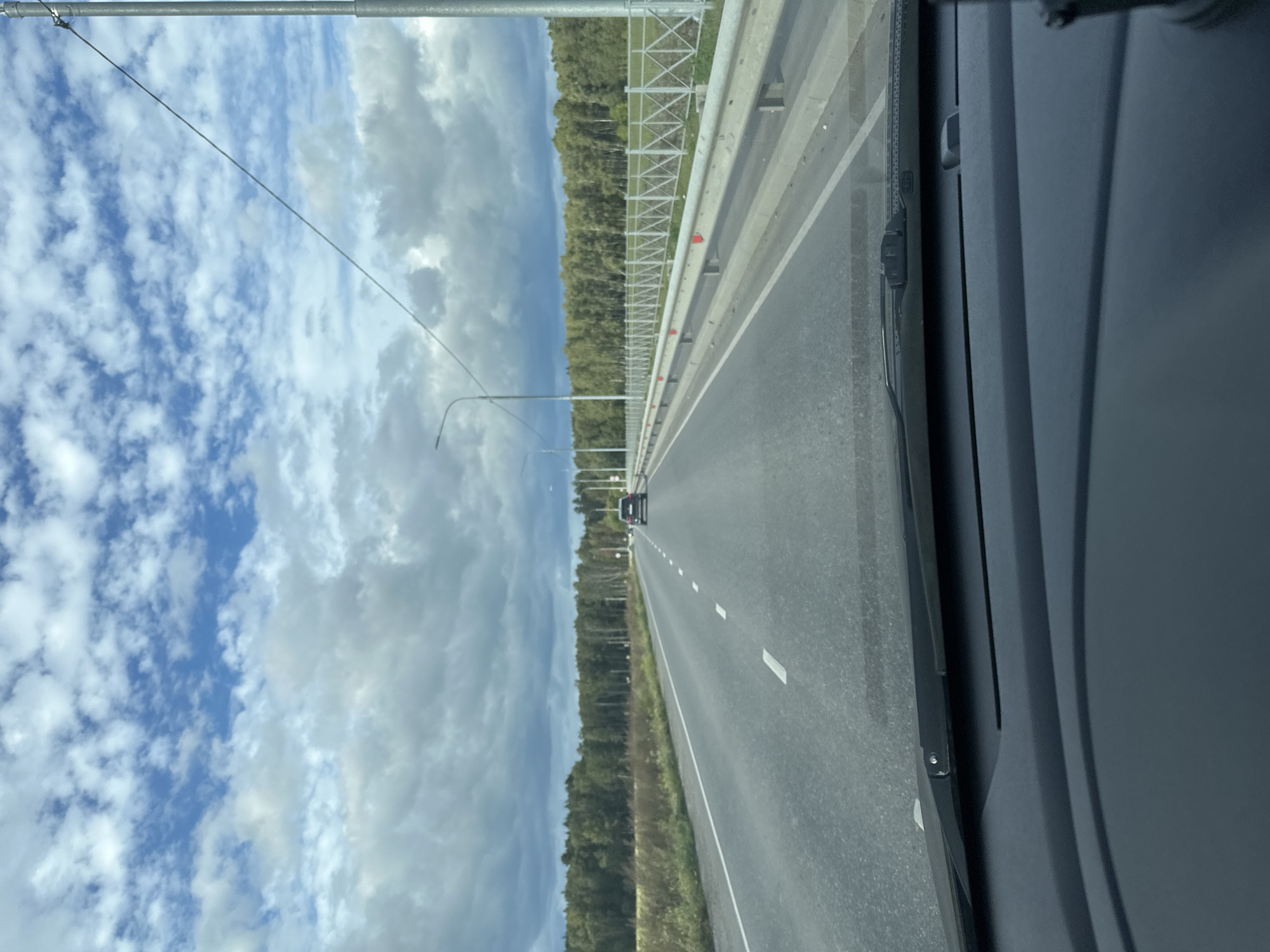 The image shows a view from inside a vehicle driving on a paved road. The road has multiple lanes and is marked with white dashed lines. To the right of the road is a metal guardrail and a white fence with a diamond pattern. Beyond the fence is a grassy area and a dense forest. In the distance, a black SUV is visible on the road. The sky above is a mix of blue sky and dramatic, heavy clouds. The dashboard of the vehicle is visible in the foreground, with the windshield taking up the upper portion of the frame. The scene appears to be in a rural or suburban area, possibly on a highway leading into or out of Lukovnya, Russia, given the presence of a forest and the road infrastructure.