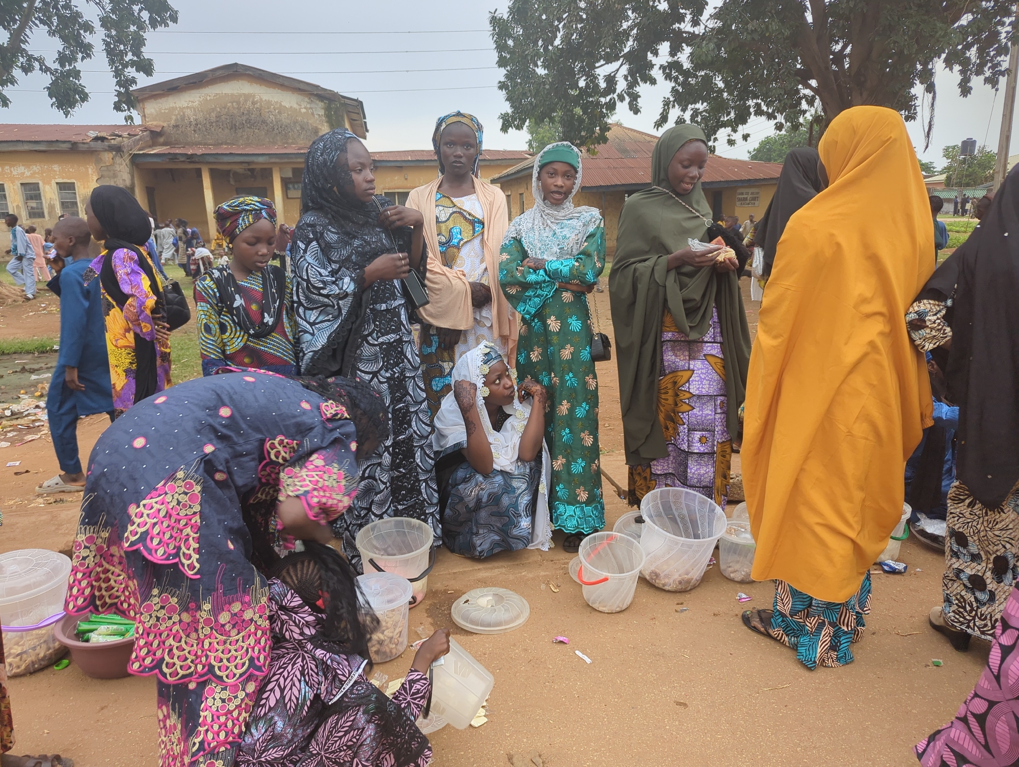 The image depicts a group of people, predominantly women and girls, gathered in an outdoor setting in what appears to be a public area in Kafanchan, Nigeria. The women and girls are dressed in a variety of colorful, traditional clothing, many wearing headscarves or hijabs. Some of the individuals are interacting with small containers and food items, suggesting they may be selling or distributing snacks. The environment appears to be somewhat dusty, with a building in the background that seems aged or in need of repair. There are also other people visible in the background, further indicating a public or gathering space.