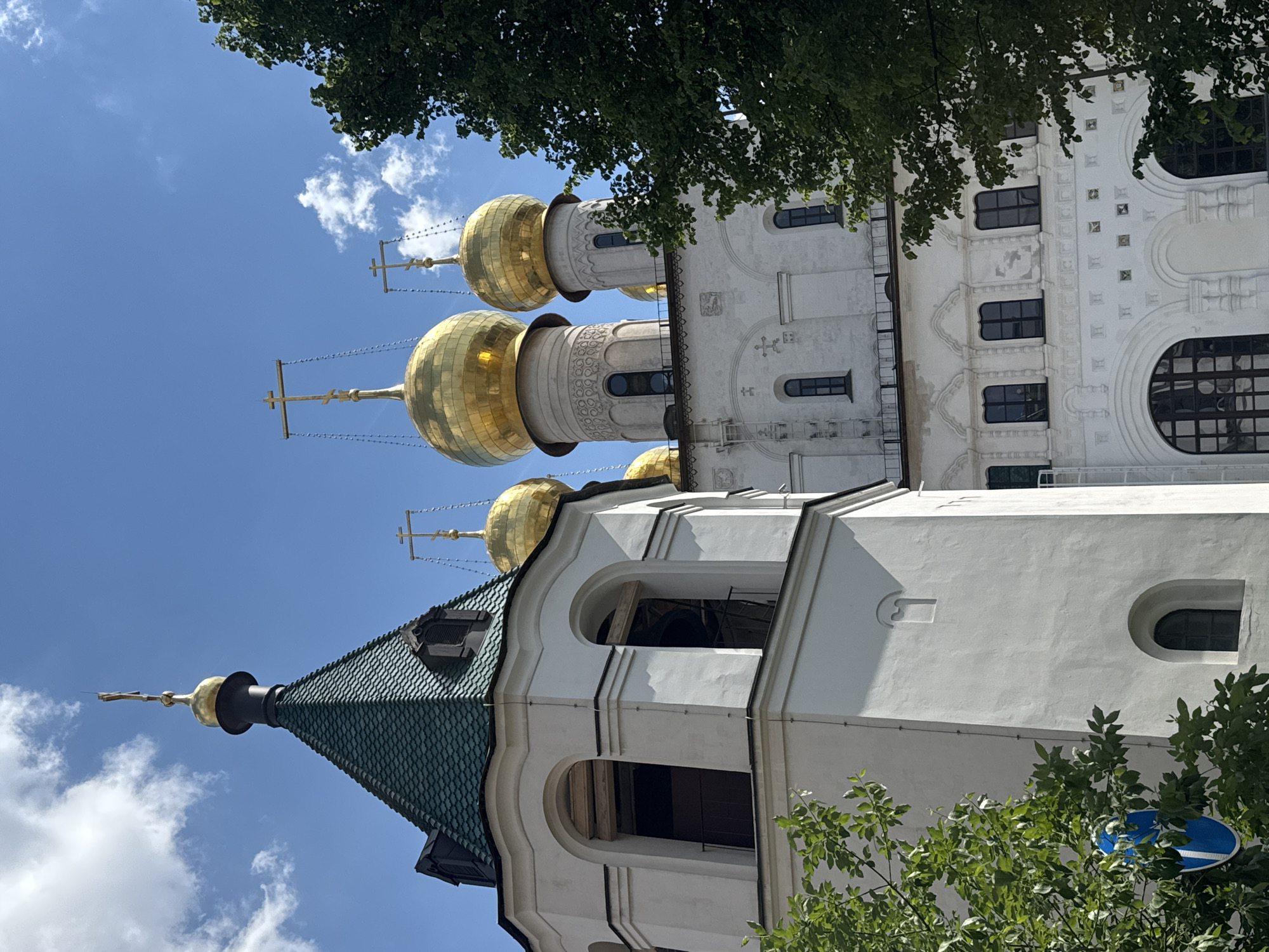 A towering white architectural structure, likely an Eastern Orthodox church or cathedral, dominates the scene, standing against a bright blue sky dotted with scattered white clouds. The building is distinguished by multiple gleaming golden onion domes, each crowned with an ornate Orthodox cross.
In the lower portion of the image, a section of the building features a dark green, possibly tiled, conical roof, which is topped with a smaller golden finial and cross. This section includes arched openings, suggesting it may be a bell tower. The main body of the church is adorned with intricate white architectural details, including various windows—some arched, some rectangular, and some with dark glass—along with decorative carvings and patterns on its facade.
Lush green foliage from nearby trees frames the top and bottom edges of the composition, partially obscuring parts of the building and sky. The lighting indicates a bright, sunny day, likely around midday. A small, circular blue and white sign is partially visible through the leaves at the bottom right. No readable text is discernible on the building or the sign.
