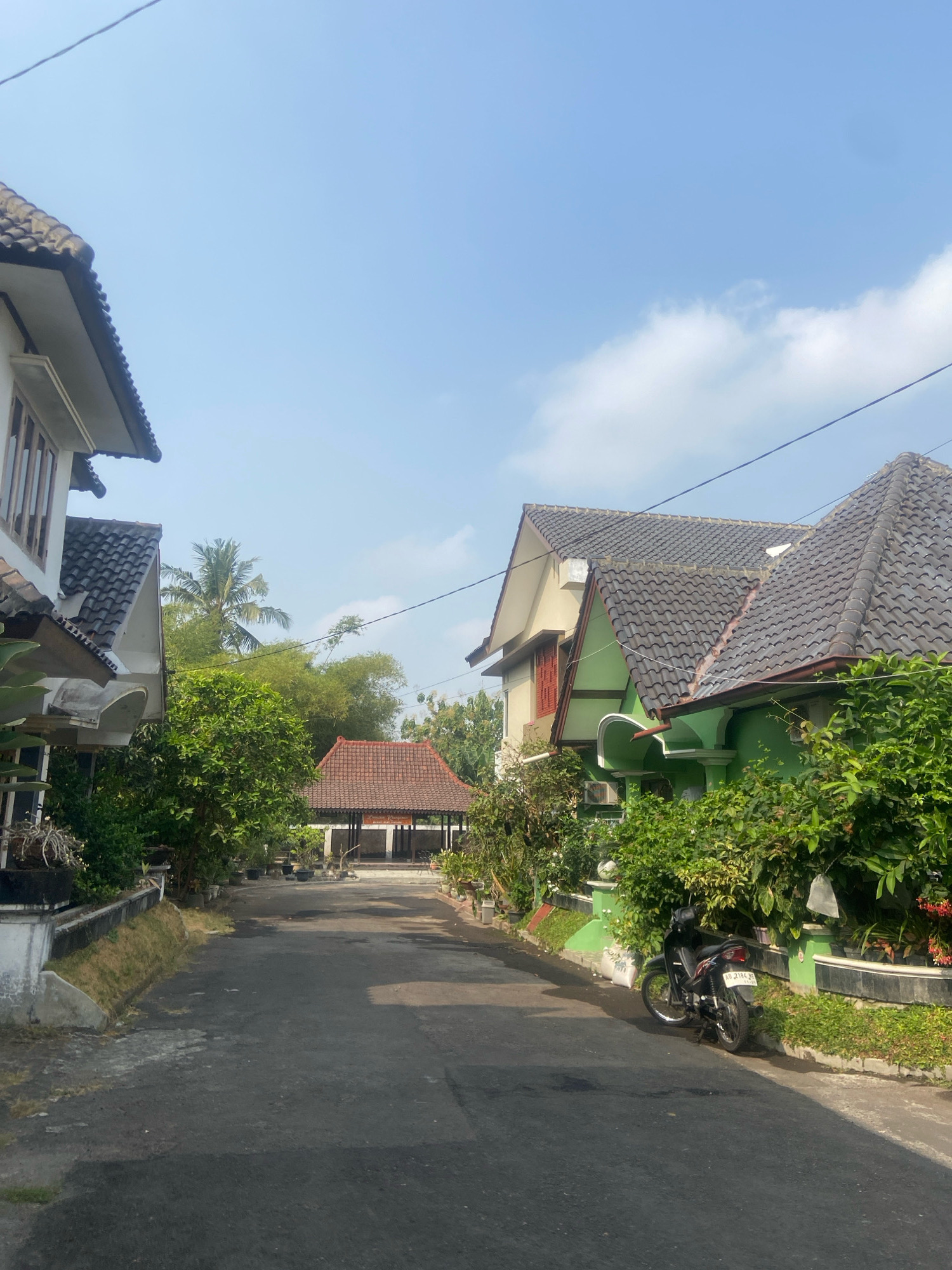 The image depicts a residential street in Sleman, Indonesia, on a clear day with a blue sky. The street is paved and appears to be of moderate width, with houses lining both sides. On the left, a house with a white facade and a gray tiled roof is partially visible, featuring windows with dark frames. Adjacent to it are lush green trees and plants.
On the right side of the street, two houses are prominently featured. The first house has a beige upper story with a brown-framed window and a dark gray tiled roof. It is connected to a second house that is painted in a vibrant green color, also with a dark gray tiled roof. This green house has a curved archway and is surrounded by dense green foliage. In front of the green house, a black motorcycle with a license plate reading "AD 2184 RE" is parked on the side of the road, with its kickstand down.
Further down the street, a traditional-looking pavilion or gazebo with a reddish-brown tiled roof and open sides is visible, set back from the road and surrounded by greenery and potted plants. The overall environment suggests a well-maintained residential area with abundant vegetation and typical Indonesian architecture. The lighting indicates it is daytime, with shadows cast on the road, suggesting the sun is to the right or slightly in front of the viewer. No people are visible in the image.