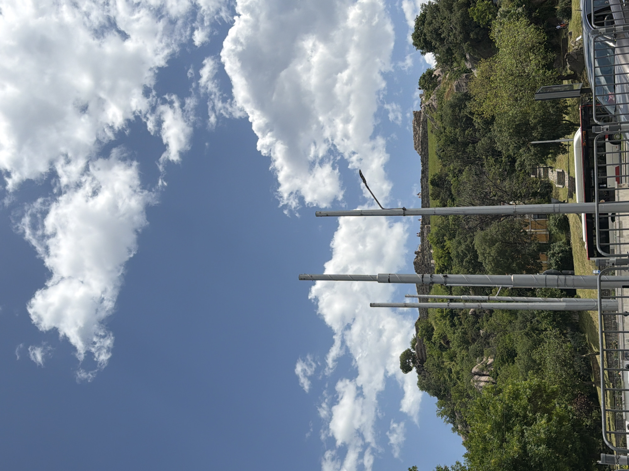 The image shows the ruins on Nebet Tepe hill in Plovdiv, Bulgaria. Several metal posts stand in the foreground. A fence and bus are visible to the right. The sky is blue with scattered clouds. The hill is covered in trees and greenery.