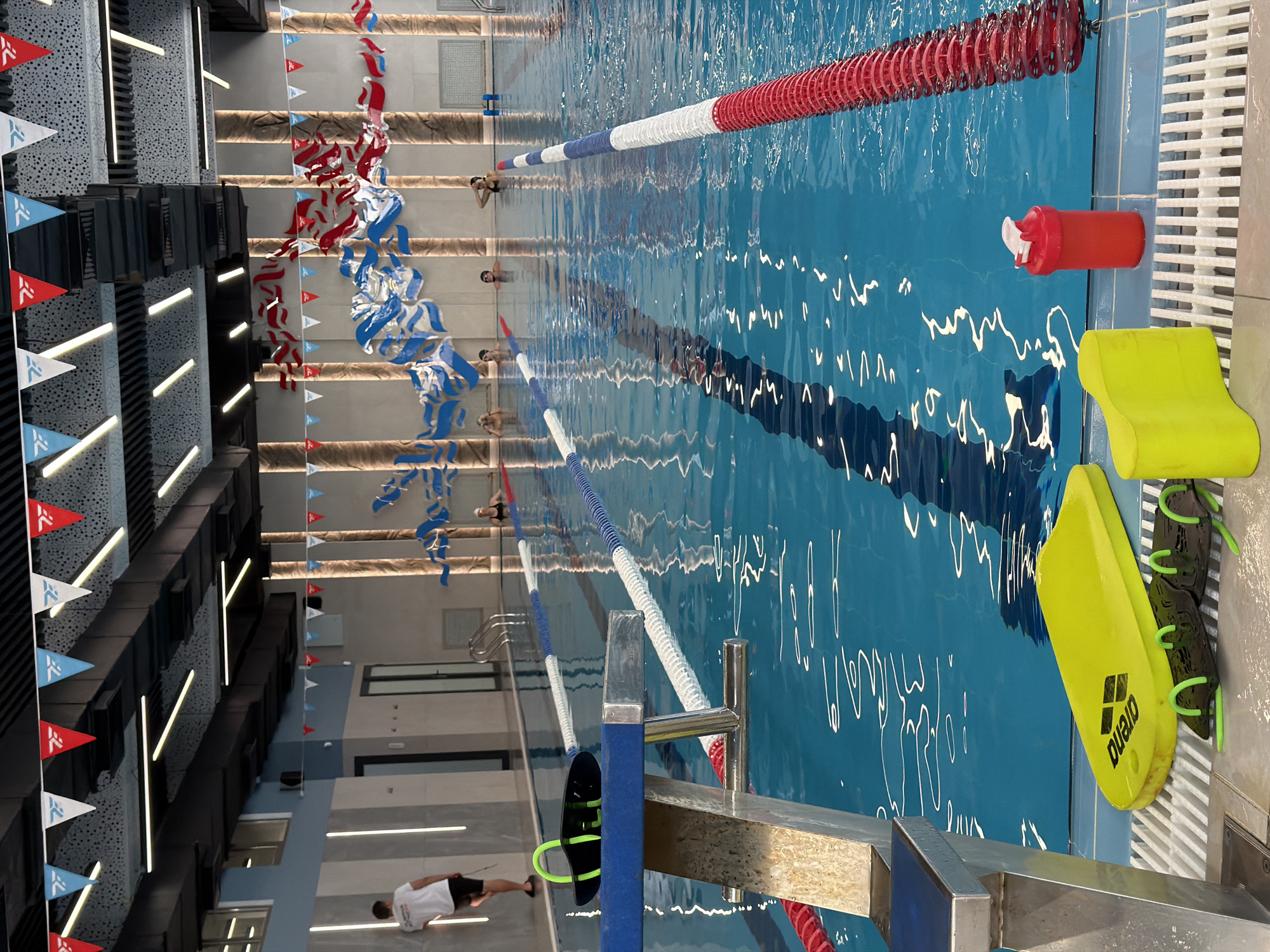 The image shows the interior of a swimming pool with multiple lanes marked by blue and red buoys. In the foreground, a blue diving platform is visible, with swimming equipment such as a yellow kickboard, black hand paddles, and a red water bottle placed on the poolside. Several people are in the pool, some swimming and others standing in the shallow end. The pool area is decorated with red, white, and blue triangular flags and wave-like fabric installations. The lighting appears to be artificial, with ceiling lights illuminating the pool area. The overall environment suggests a public or training facility for swimming. There is no visual information to suggest this location is in Bishkek City, Kyrgyzstan.